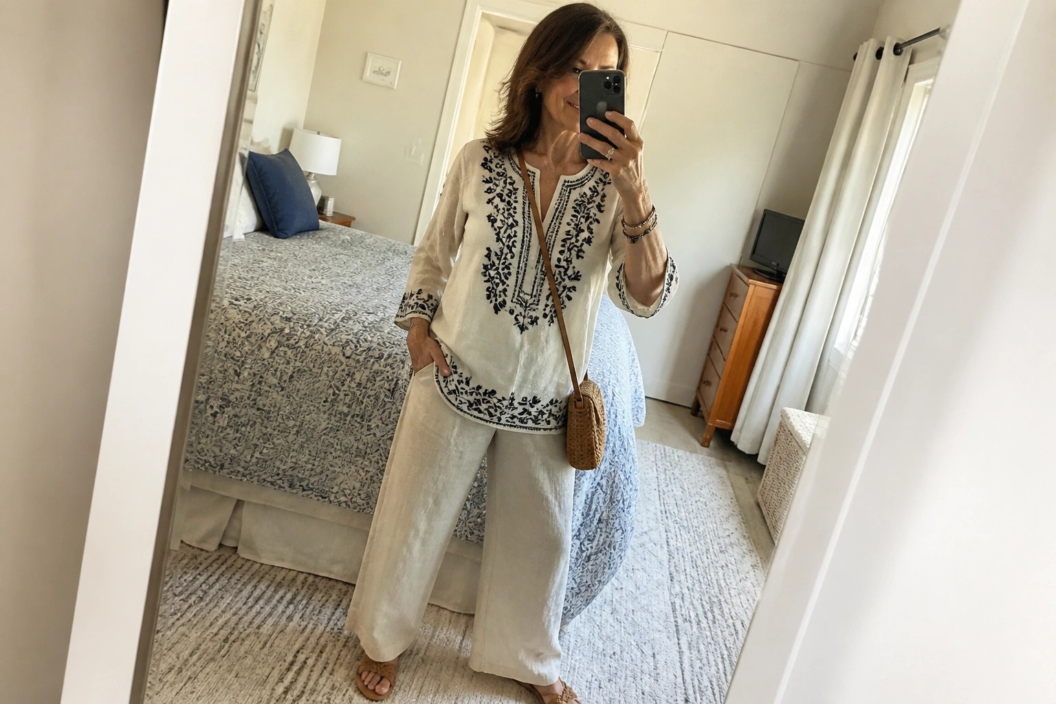 Woman in white embroidered tunic and wide-leg pants taking bedroom mirror selfie