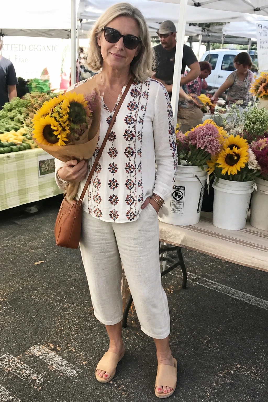 Blonde woman in embroidered white top holding sunflowers at farmers market.