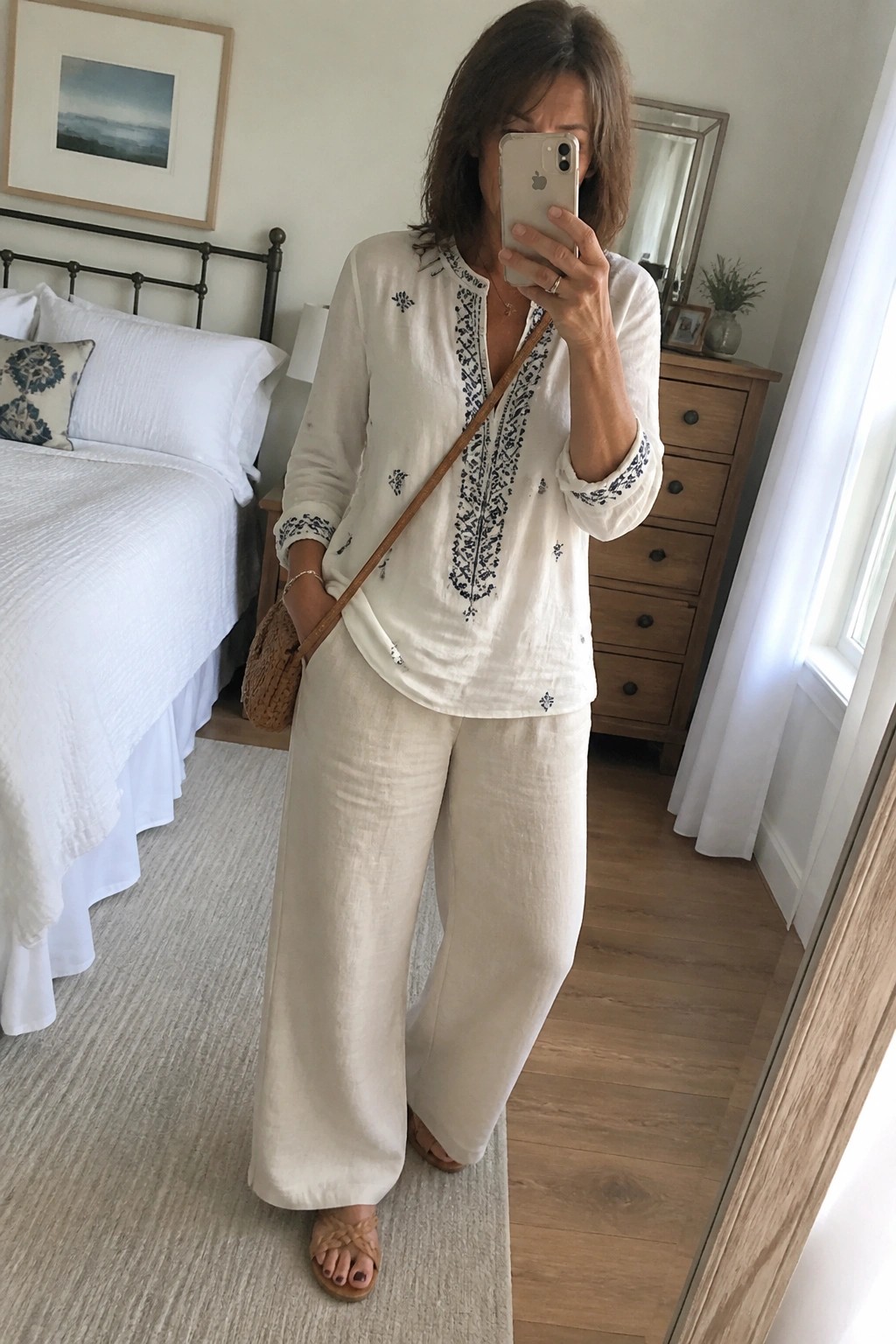 Woman in white embroidered tunic and wide-leg pants taking bedroom mirror selfie