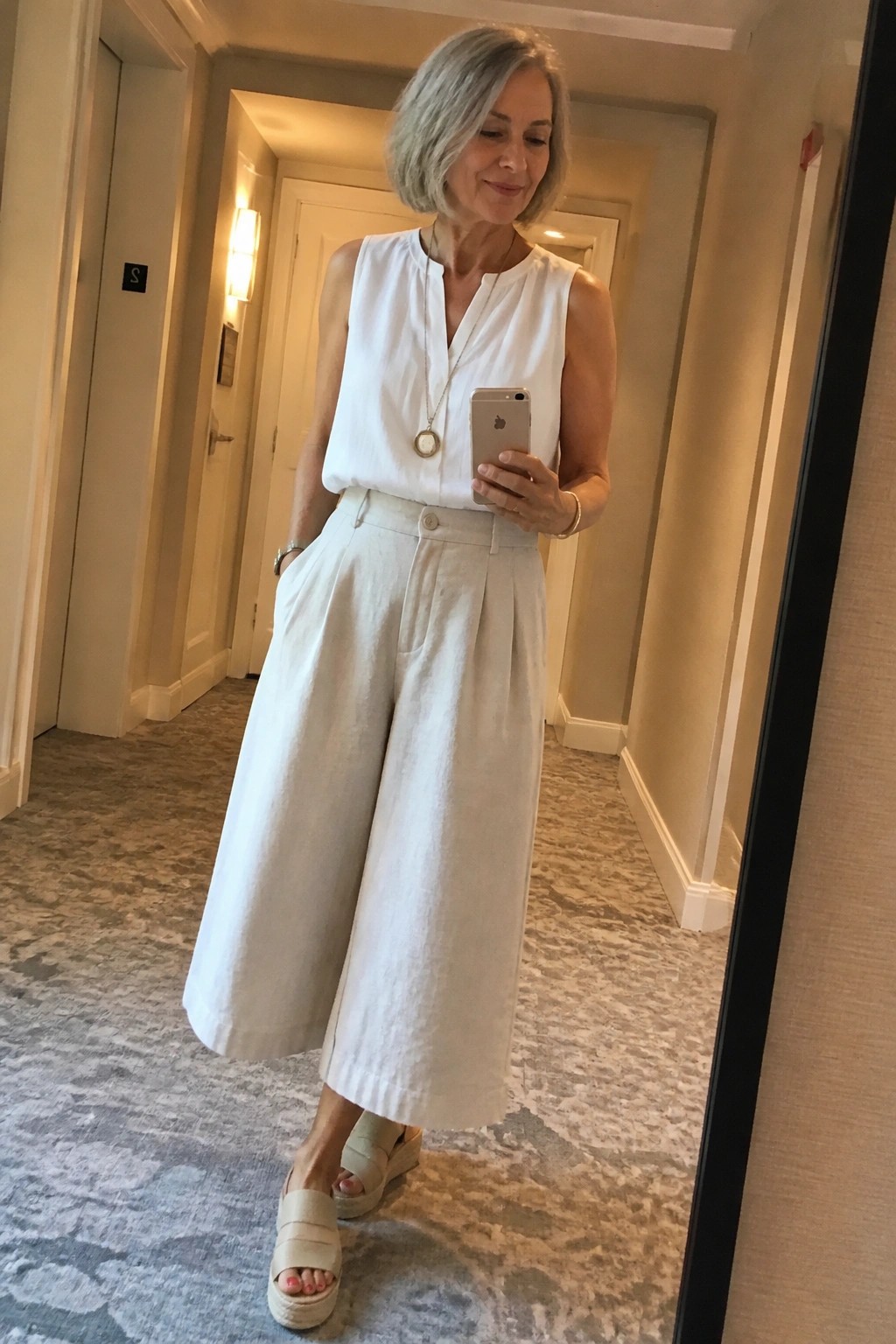 Woman with short gray hair in white top and wide-leg pants taking mirror selfie