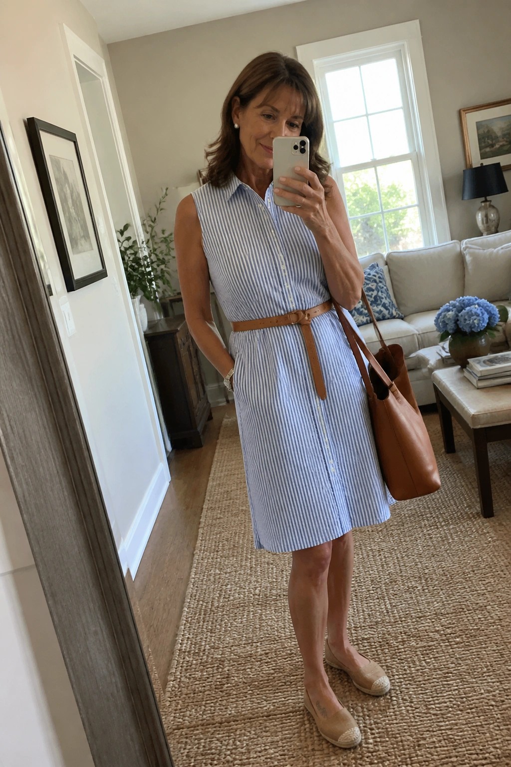 Woman in blue striped sleeveless dress holding phone and brown leather tote in living room
