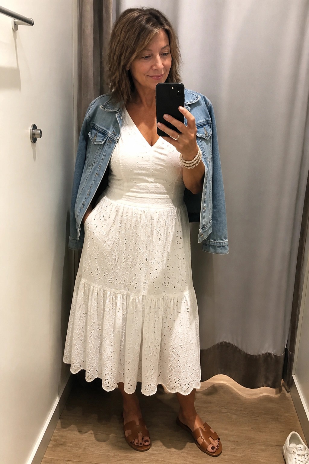 Woman in white eyelet dress and denim jacket taking mirror selfie in fitting room