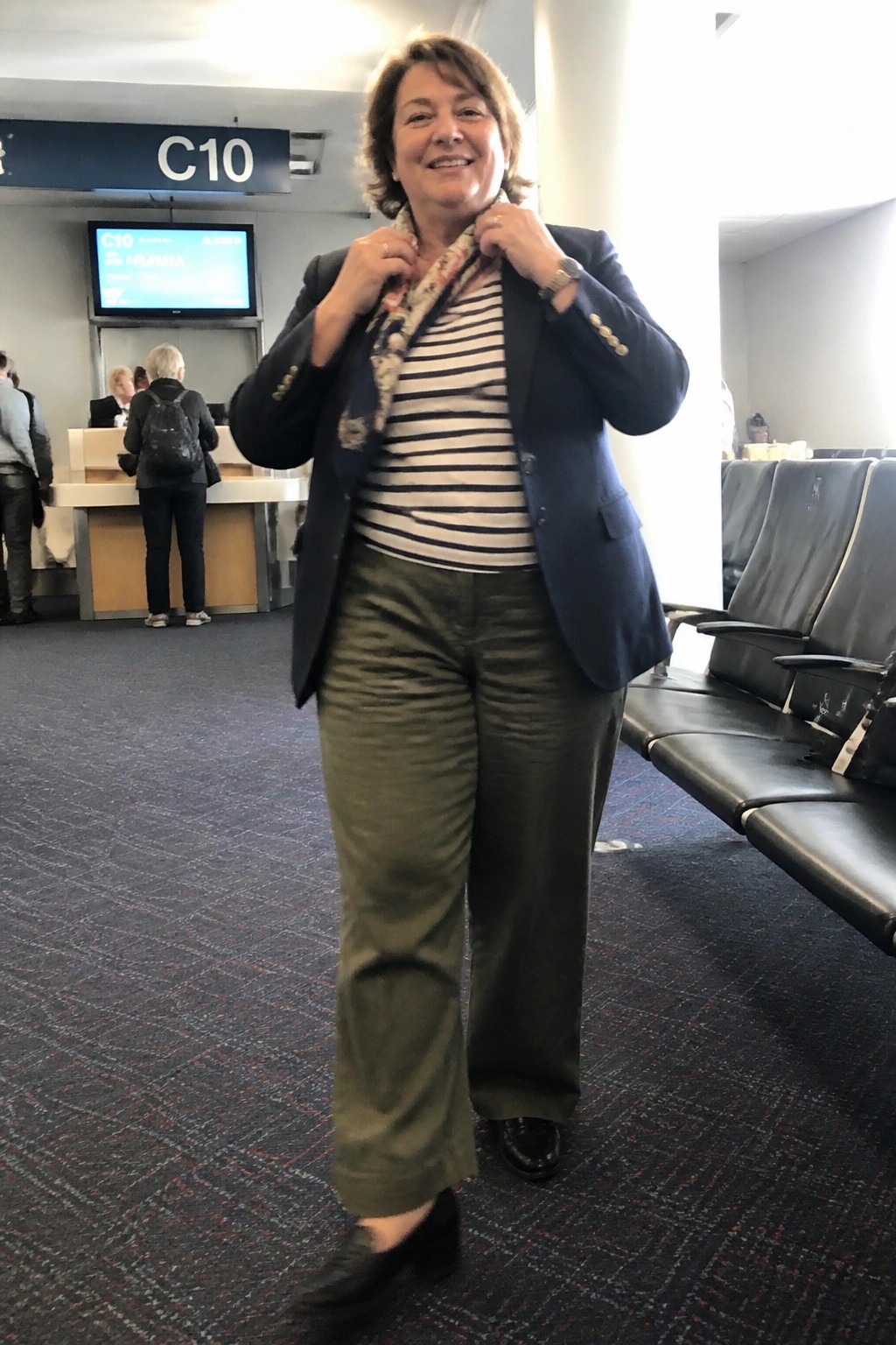 Woman in navy blazer and olive pants adjusting scarf at airport gate C10