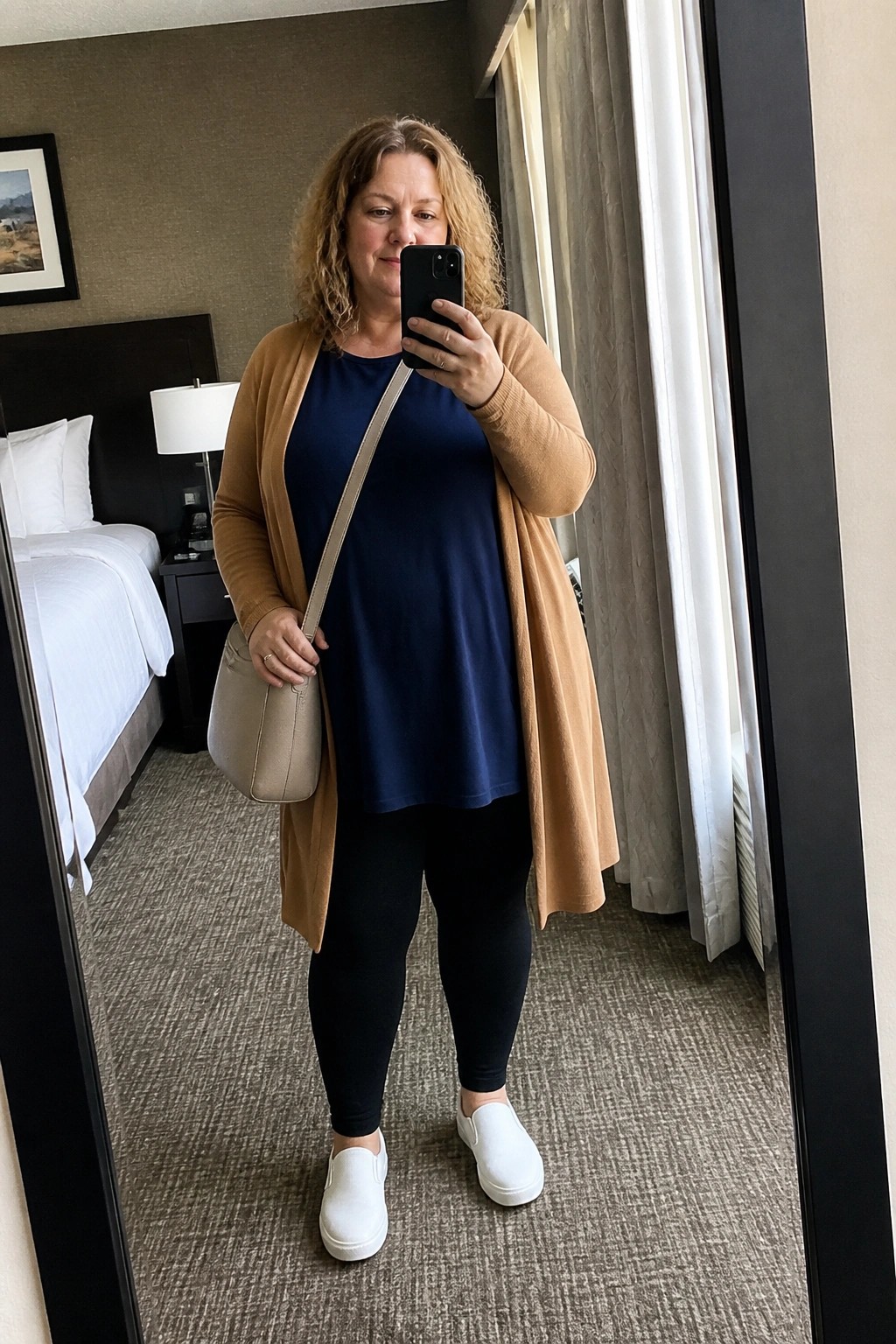 Woman in blue top and tan cardigan taking mirror selfie in hotel room