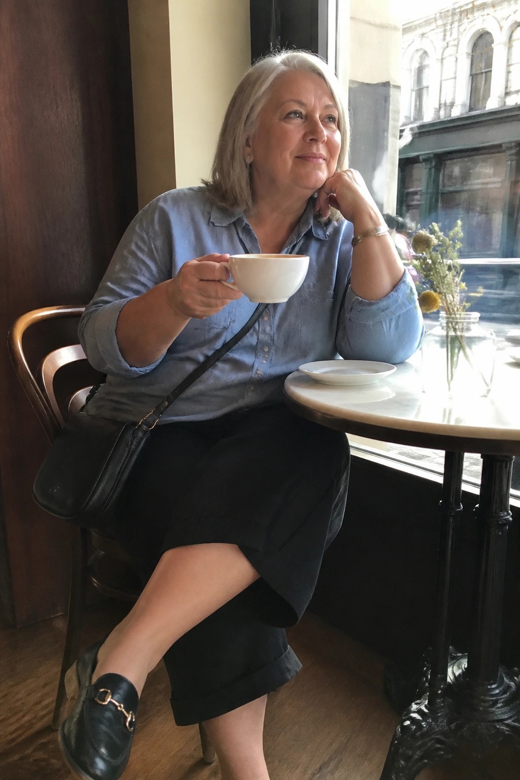 Gray-haired woman in blue shirt holds white cup at marble cafe table