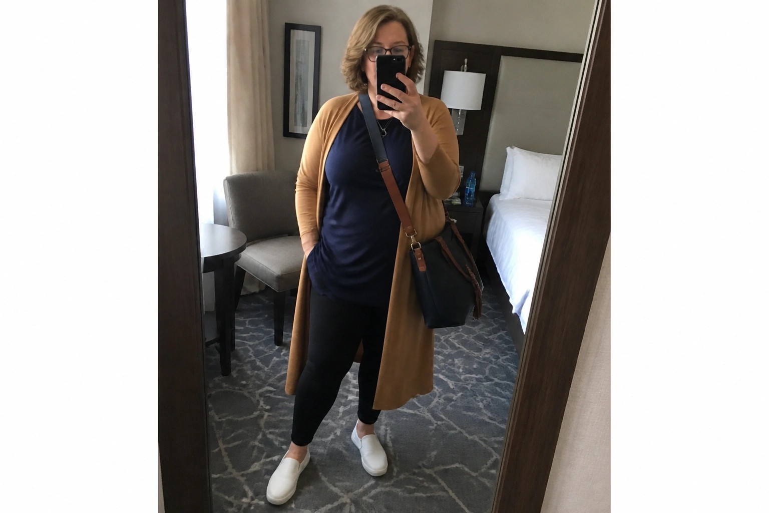 Woman in blue top and tan cardigan taking mirror selfie in hotel room