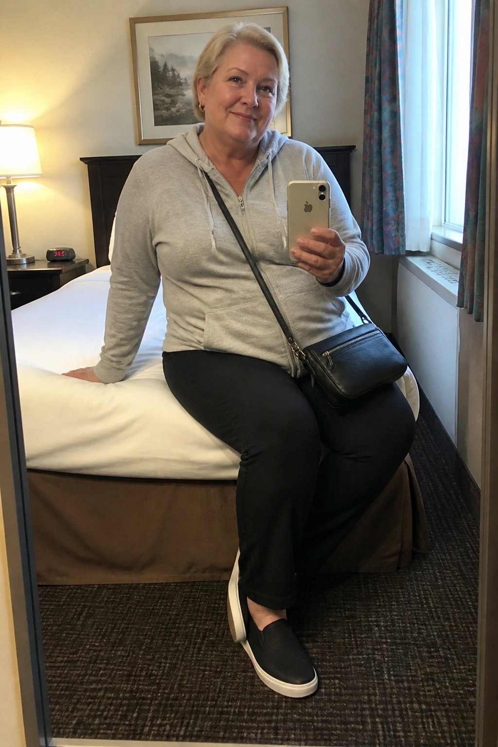 Blonde woman in gray hoodie taking mirror selfie on hotel bed with iPhone