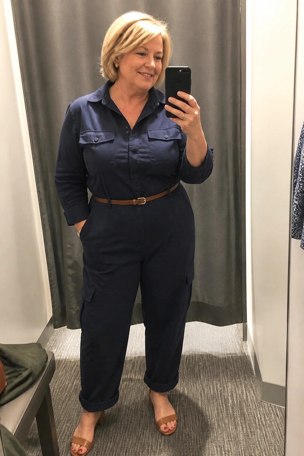 Plus size woman wearing a navy blue utility jumpsuit with brown belt and brown sandals while taking a mirror selfie in a fitting room.
