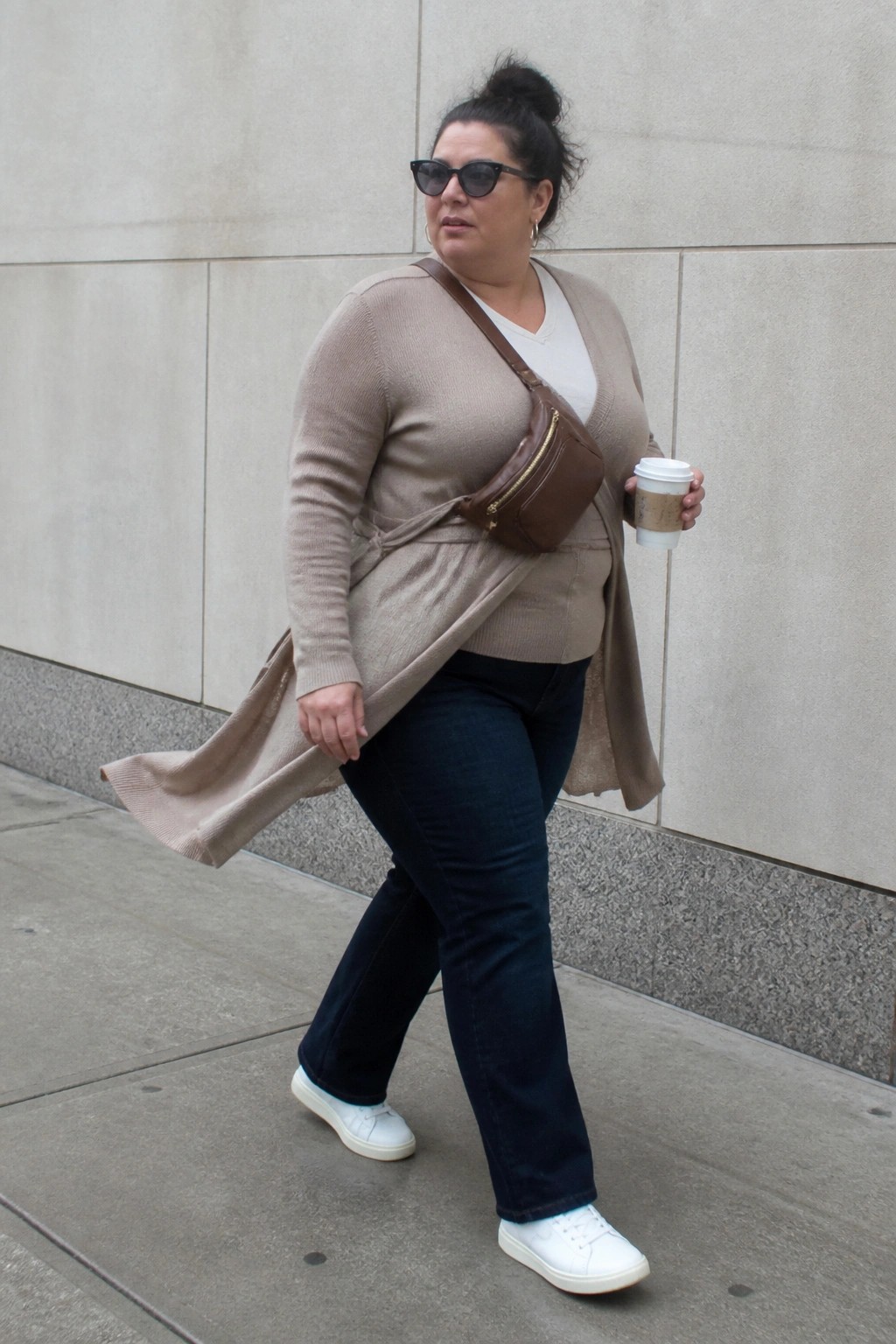 Woman in beige cardigan and jeans walks with coffee and brown bag