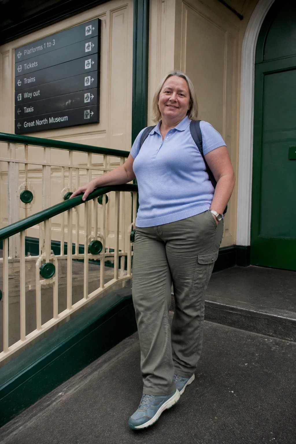 Woman in blue polo shirt and green pants leans on railing by station sign.