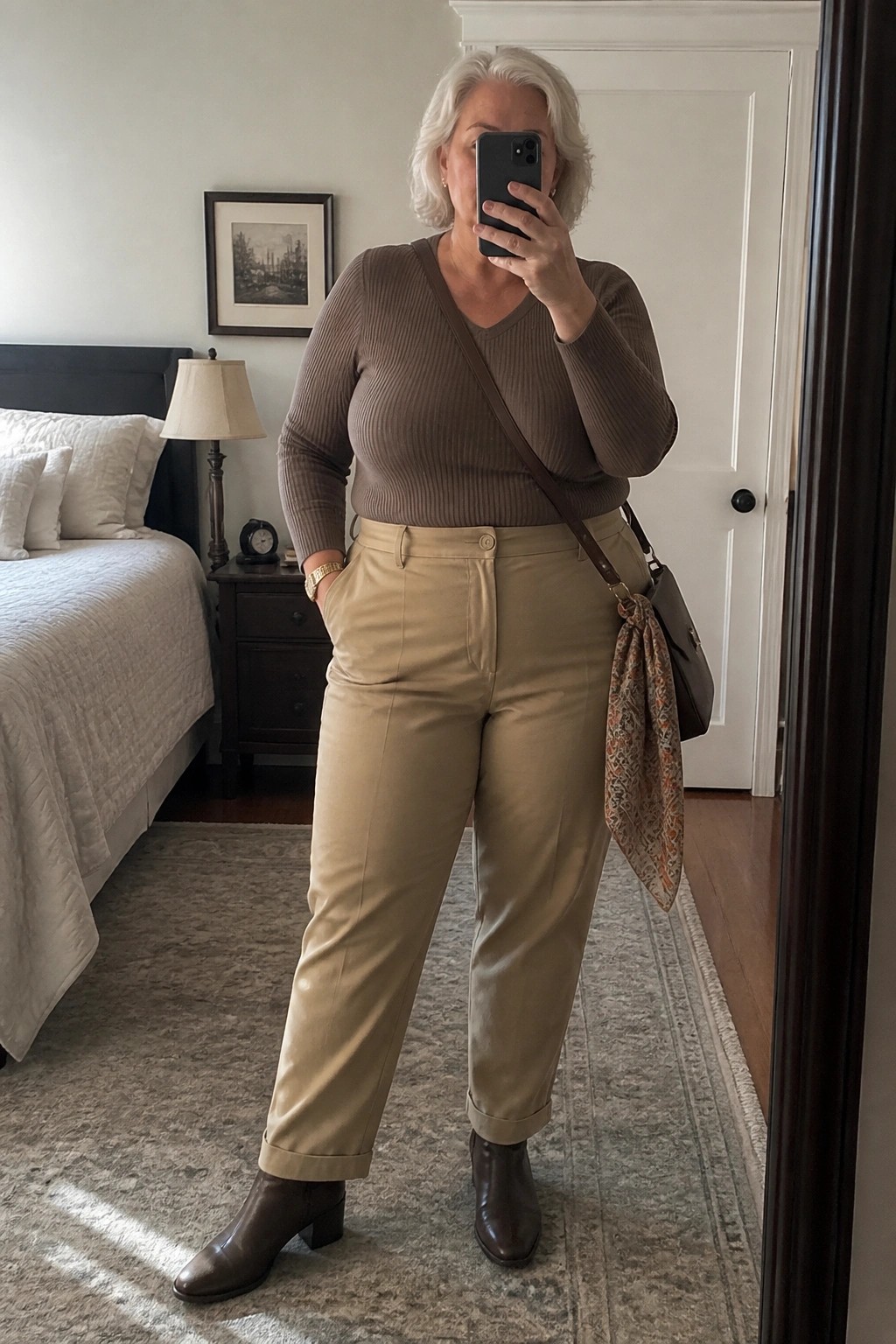 A plus size woman with short white hair wearing a brown ribbed V-neck sweater, beige pants, brown ankle boots, and carrying a brown crossbody bag with a patterned scarf, standing in a bedroom taking a mirror selfie.