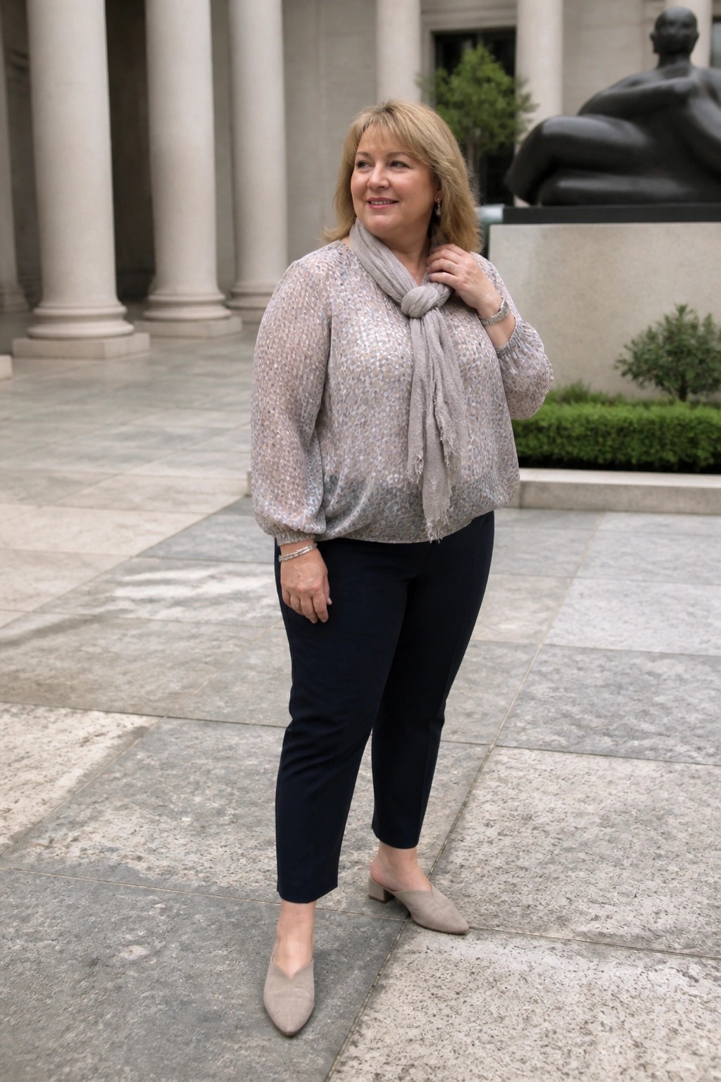 Woman in patterned blouse with gray scarf stands before columns and sculpture.