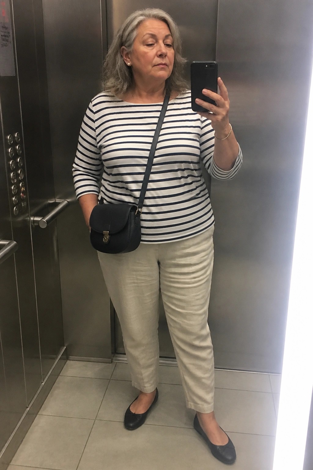 Woman with gray hair in striped shirt and beige pants taking selfie in elevator