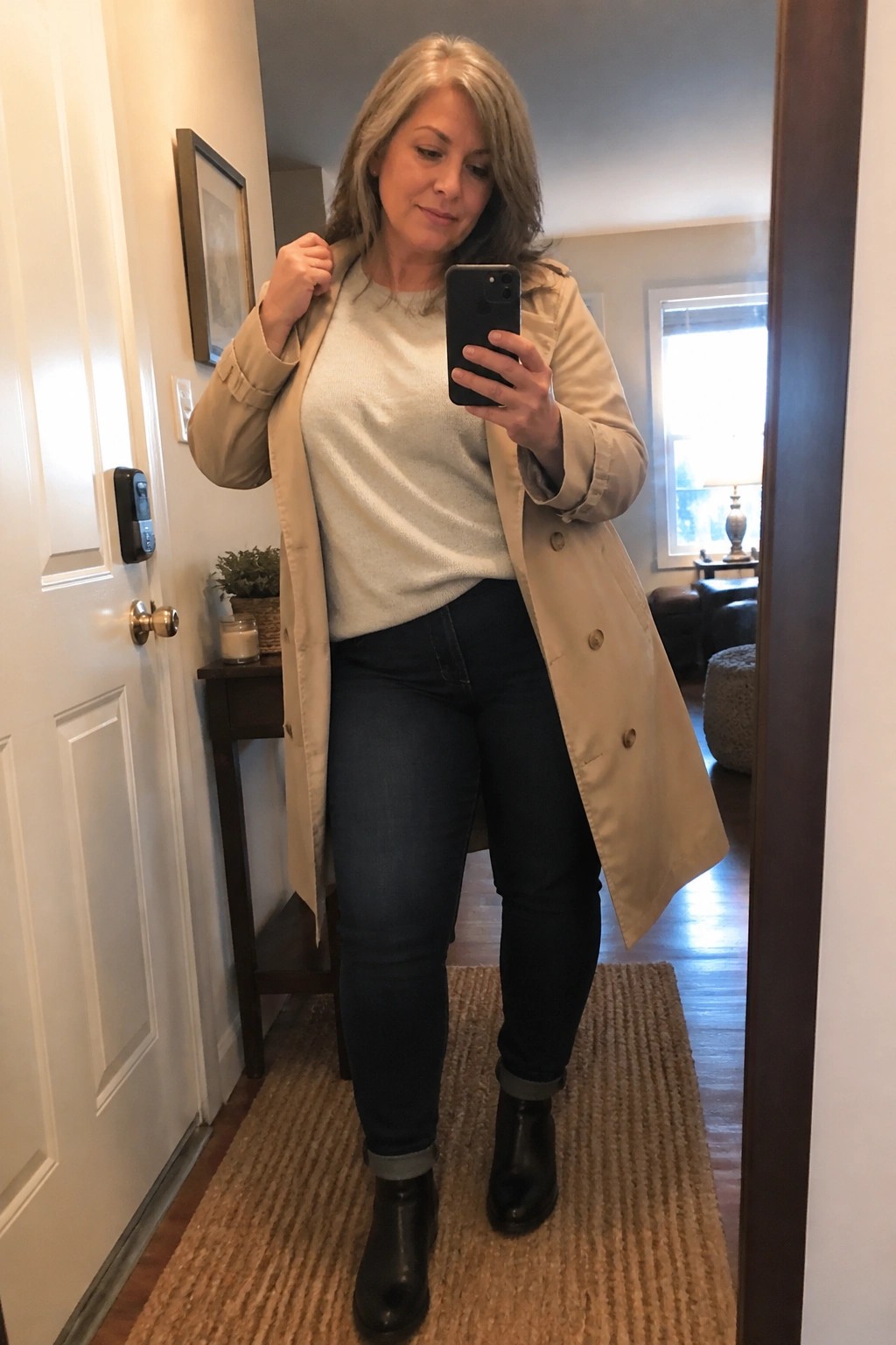 Gray-haired woman in beige trench coat taking mirror selfie in hallway.