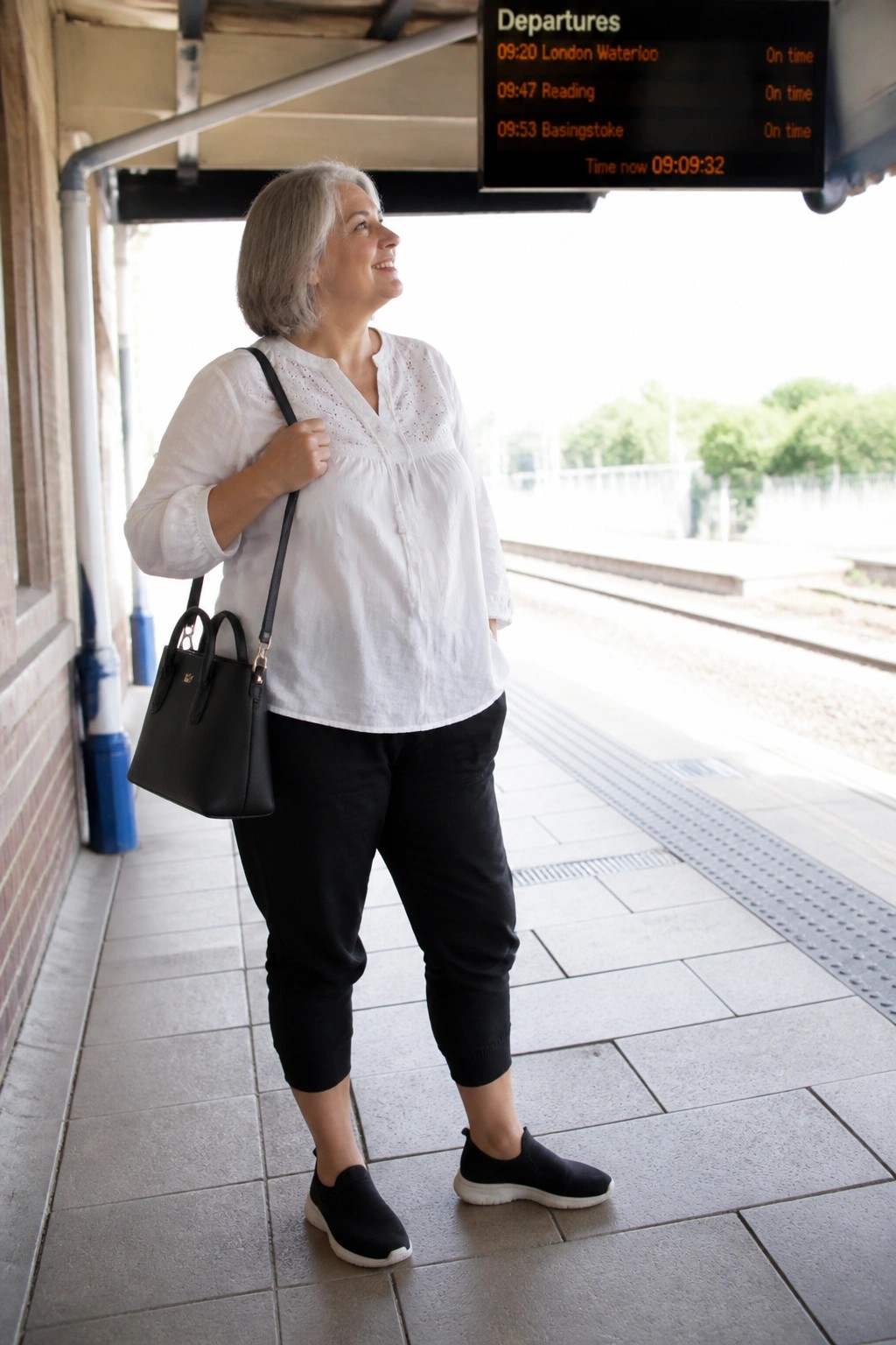 Plus size woman over 50 wearing a white eyelet blouse tucked into cropped black pants with black slip on sneakers and carrying a black handbag on a train platform.