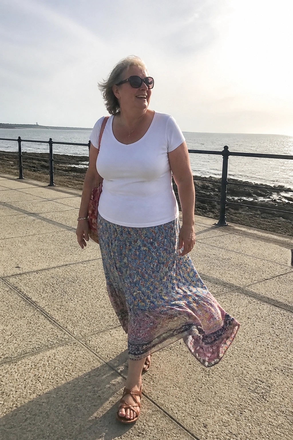 Woman wearing white scoop neck t-shirt tucked into long floral maxi skirt with blue and pink prints plus brown strappy sandals and shoulder bag