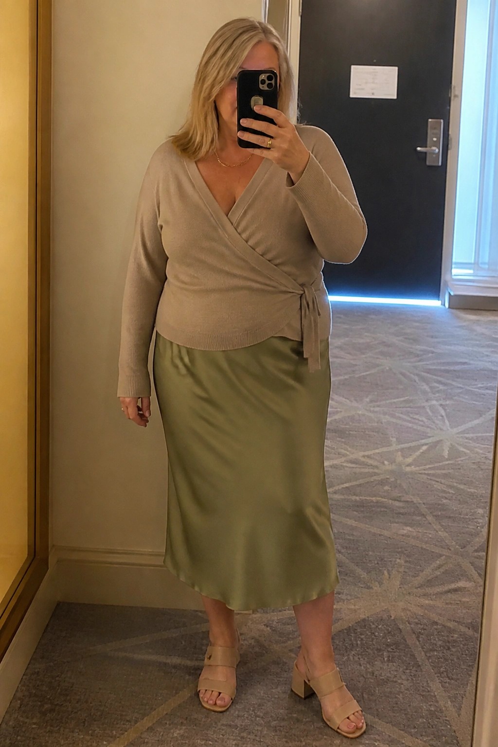 Blonde woman in beige wrap sweater and green satin skirt taking mirror selfie