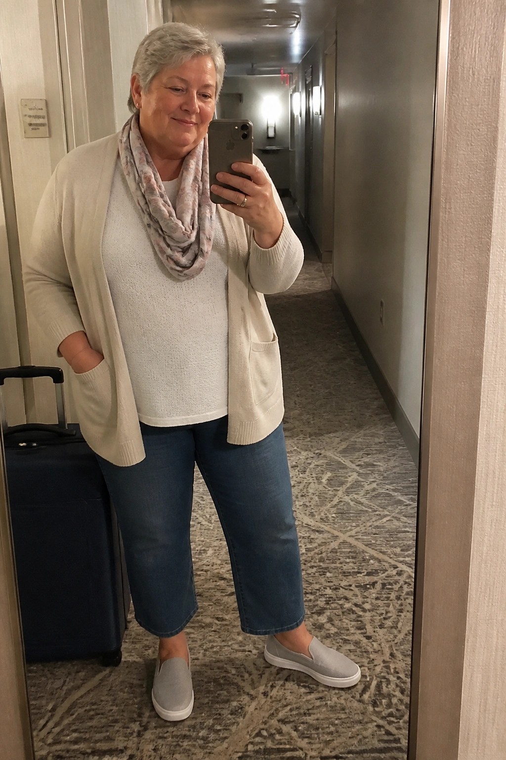 Woman in cream cardigan and jeans takes mirror selfie in hotel hallway beside blue suitcase