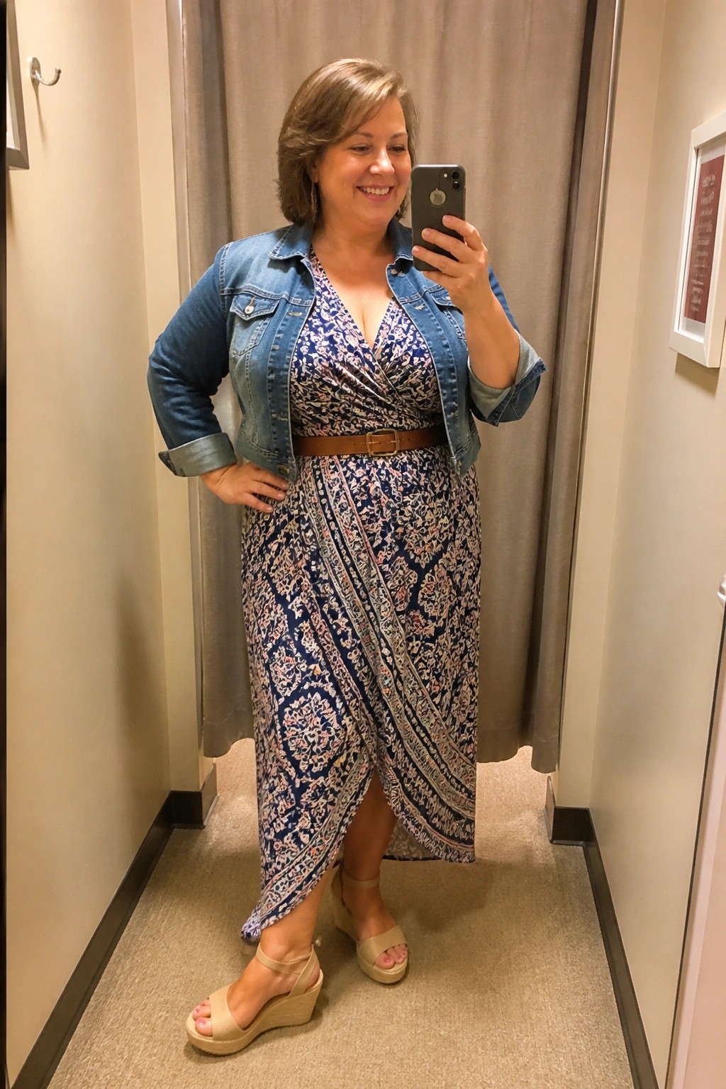 Woman in denim jacket and patterned wrap dress taking mirror selfie in fitting room