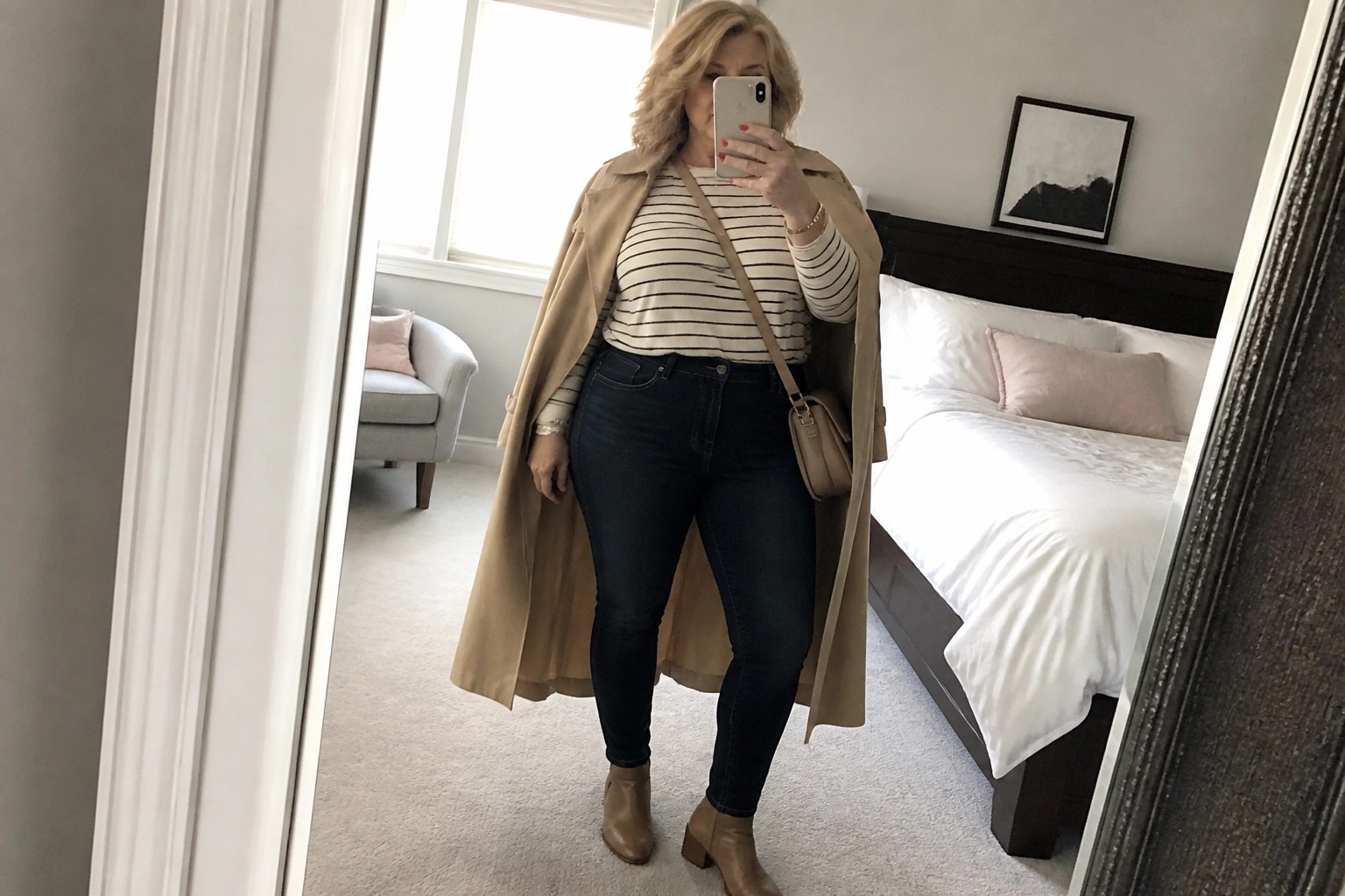 Woman in beige trench coat over striped shirt and jeans taking bedroom mirror selfie.
