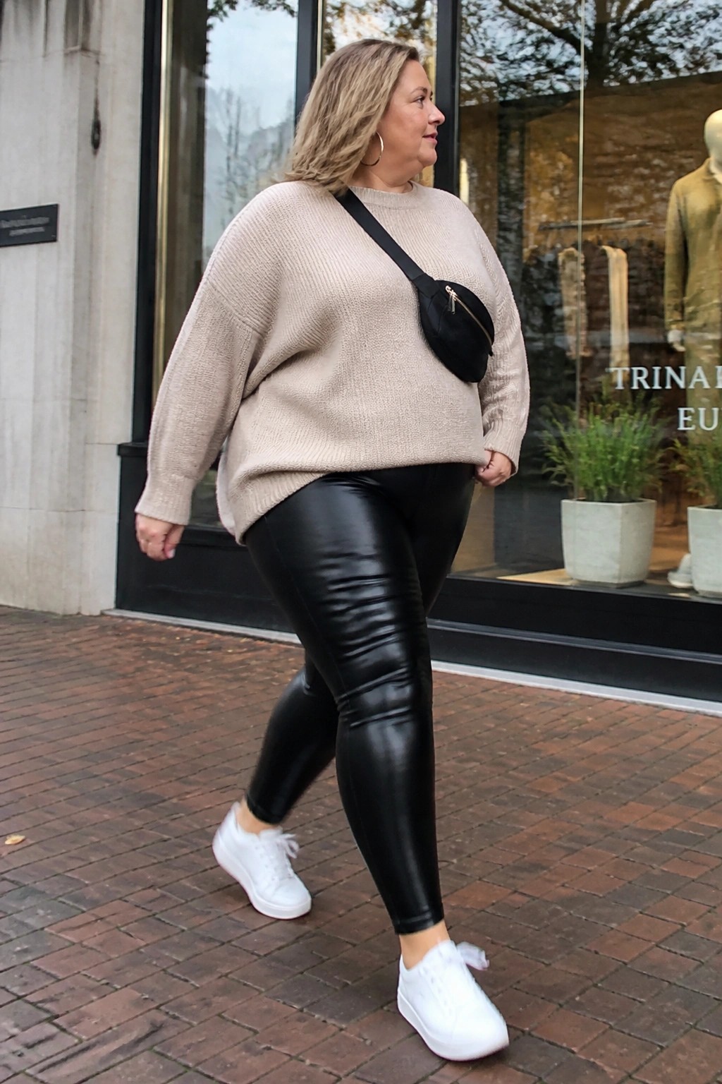 Woman in beige sweater and black leggings walking past storefront on brick sidewalk