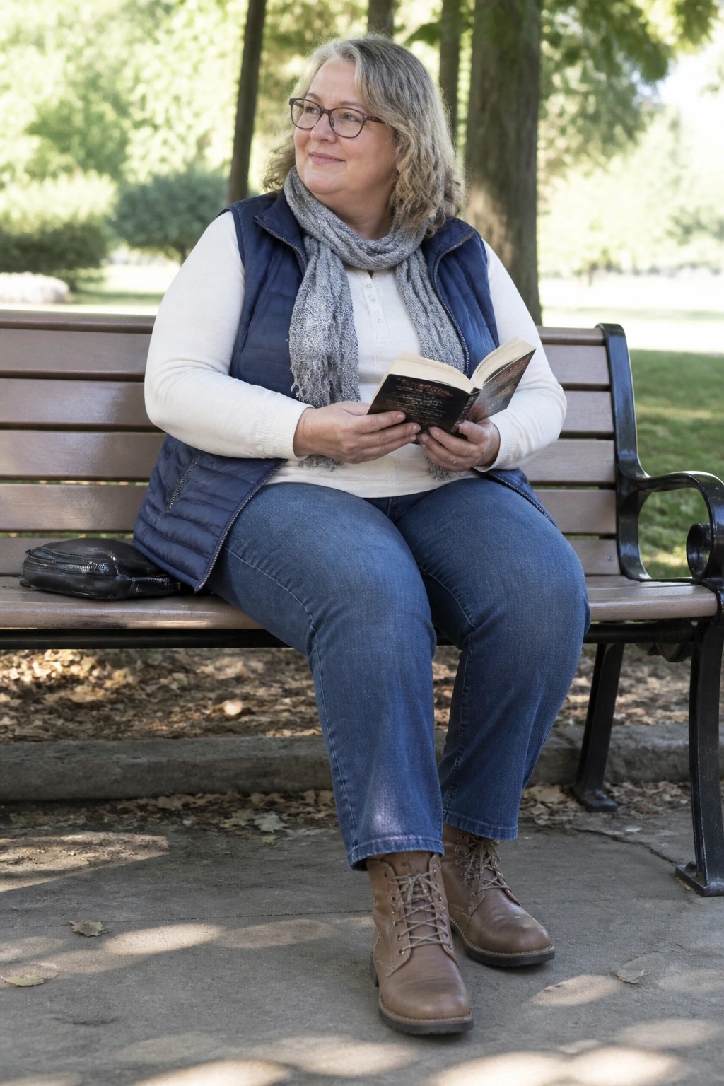 A plus size woman over 50 on a bench in a navy puffer vest over a white shirt with a gray scarf blue jeans and brown lace up boots holding an open book.