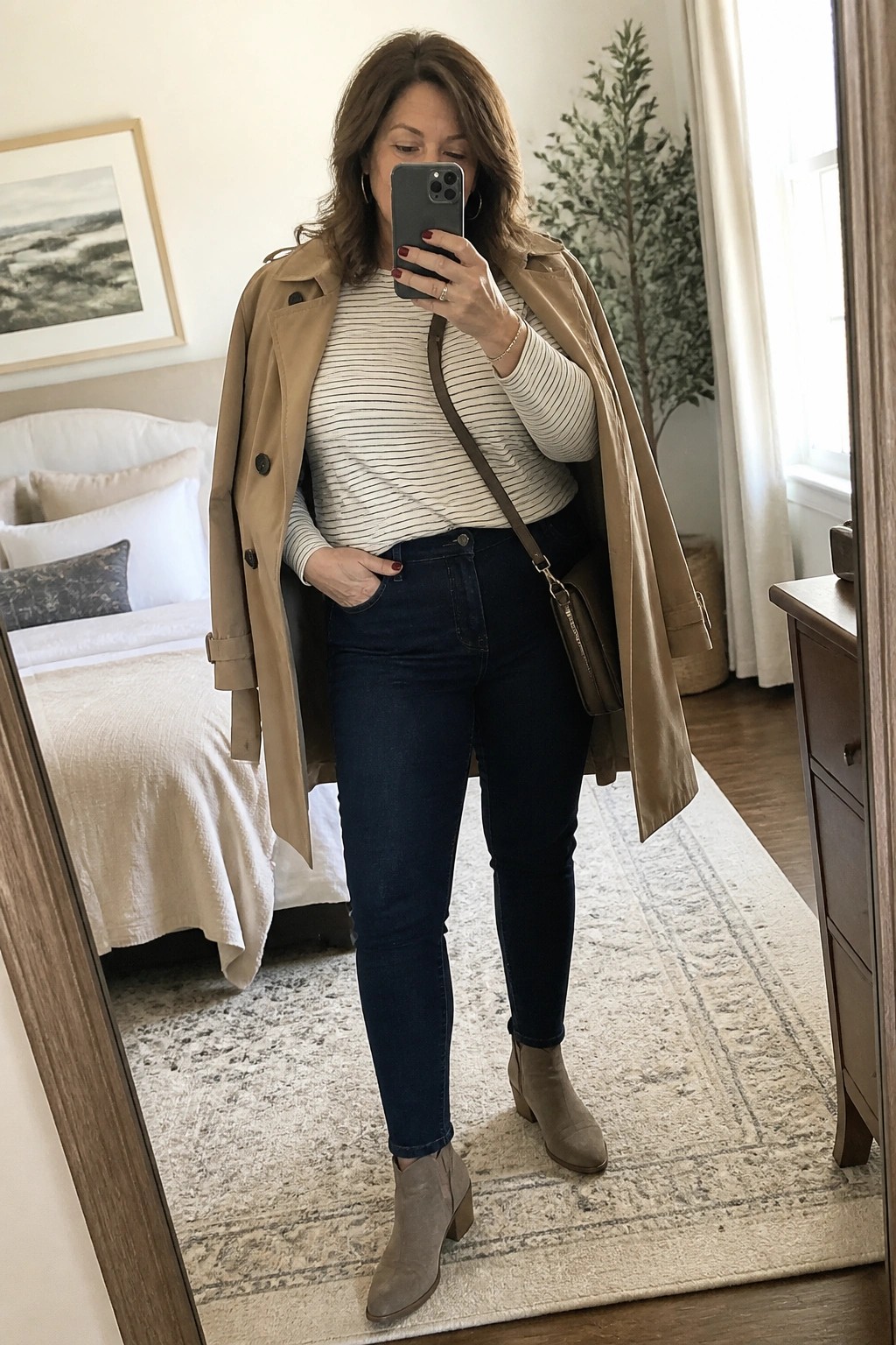 Woman in beige trench coat over striped shirt and jeans taking bedroom mirror selfie.