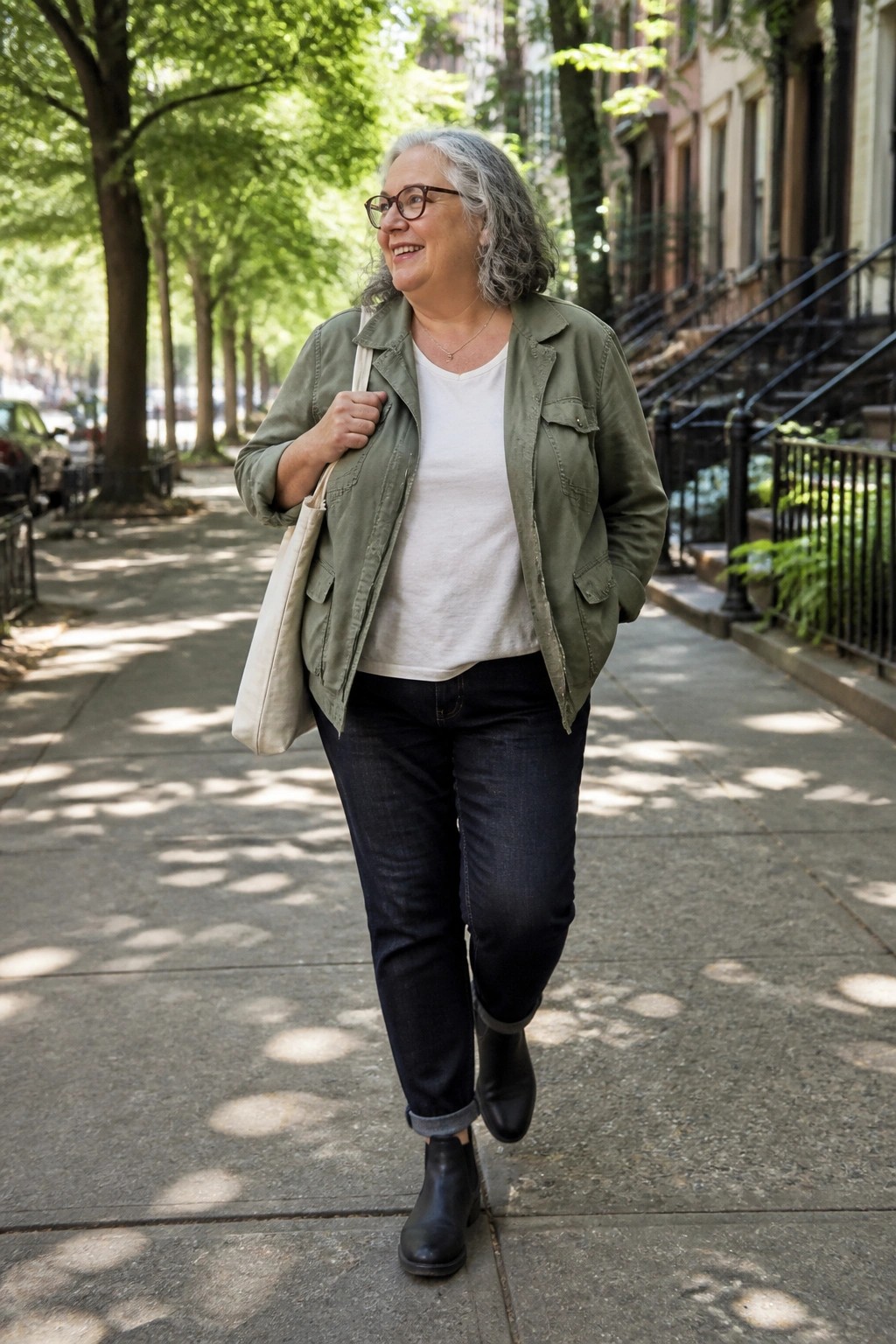 Smiling gray-haired woman in green jacket walks sunny tree-lined city sidewalk.