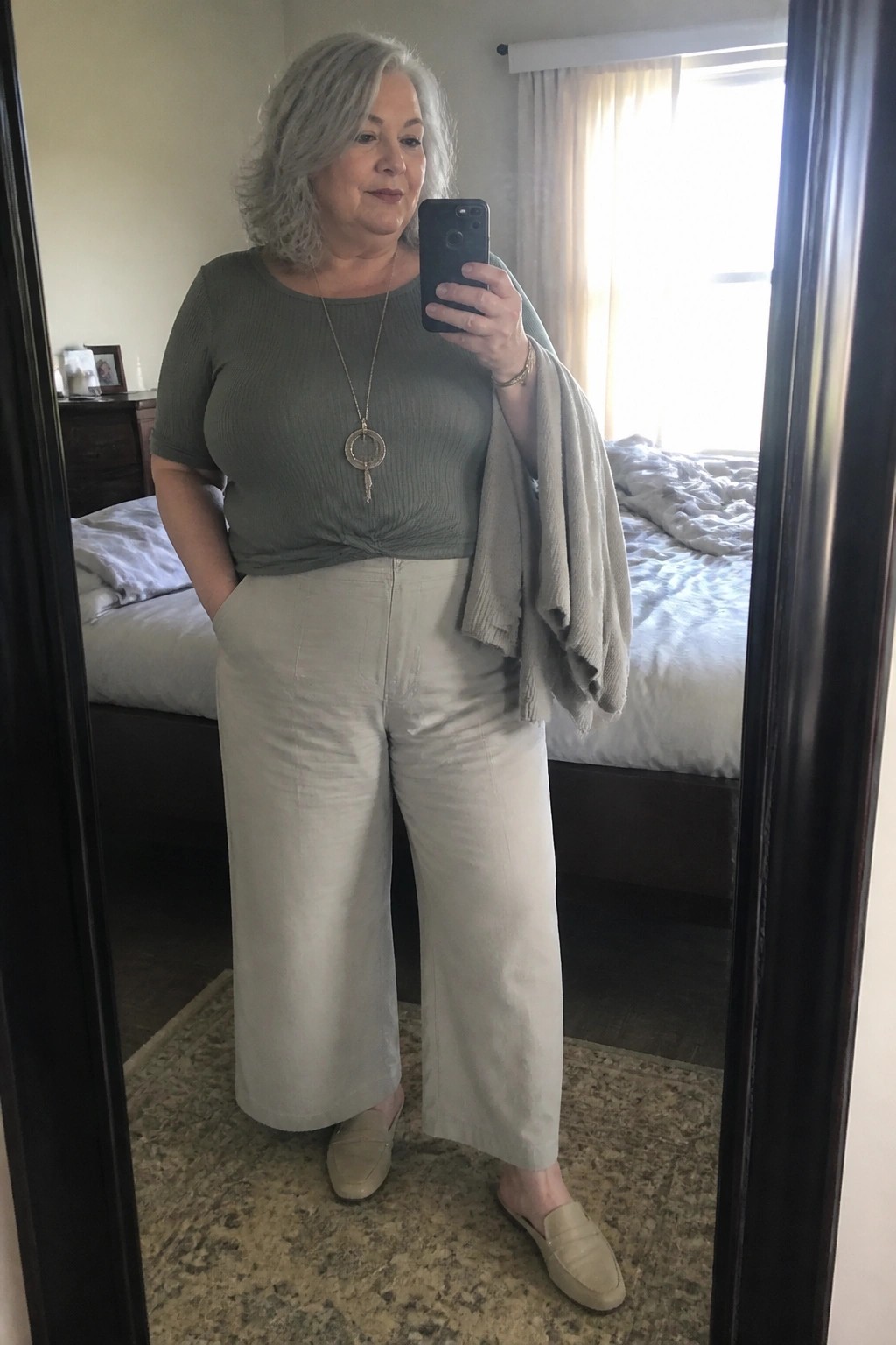 Plus size woman over 50 wearing a sage green ribbed knit top knotted at the waist with wide leg beige pants, a beige cardigan draped over one shoulder, beige loafers, and a long pendant necklace while taking a mirror selfie.