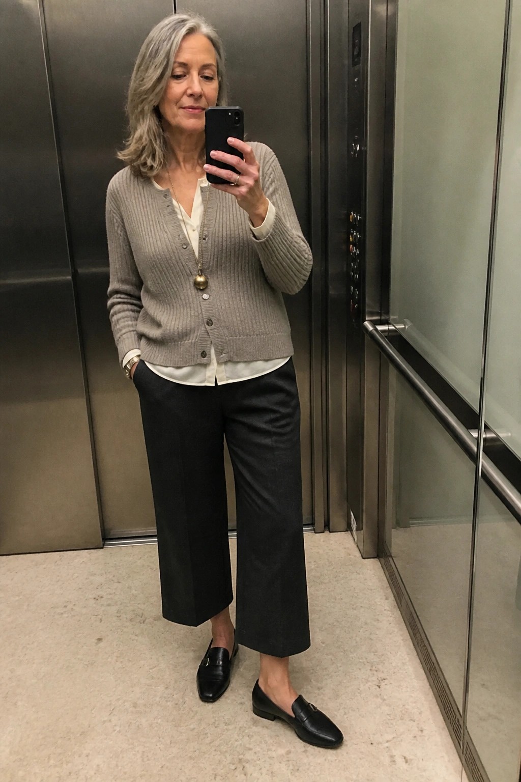 Gray-haired woman in beige cardigan and black pants taking elevator selfie