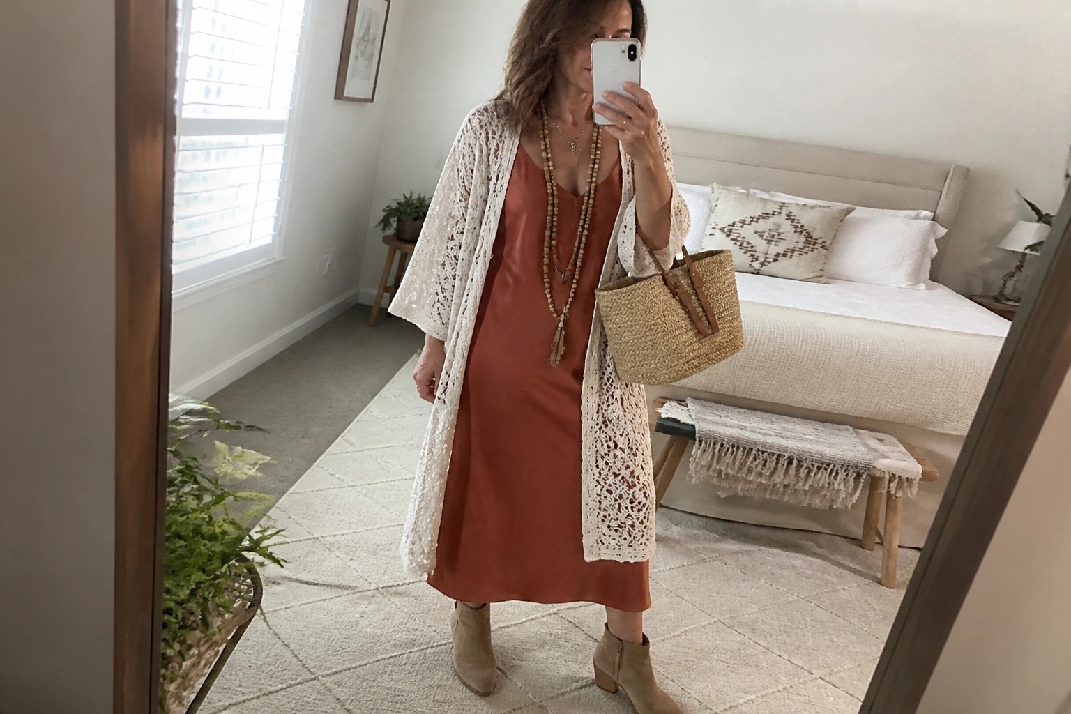 Woman in rust dress and cream open-knit cardigan taking bedroom mirror selfie with woven bag