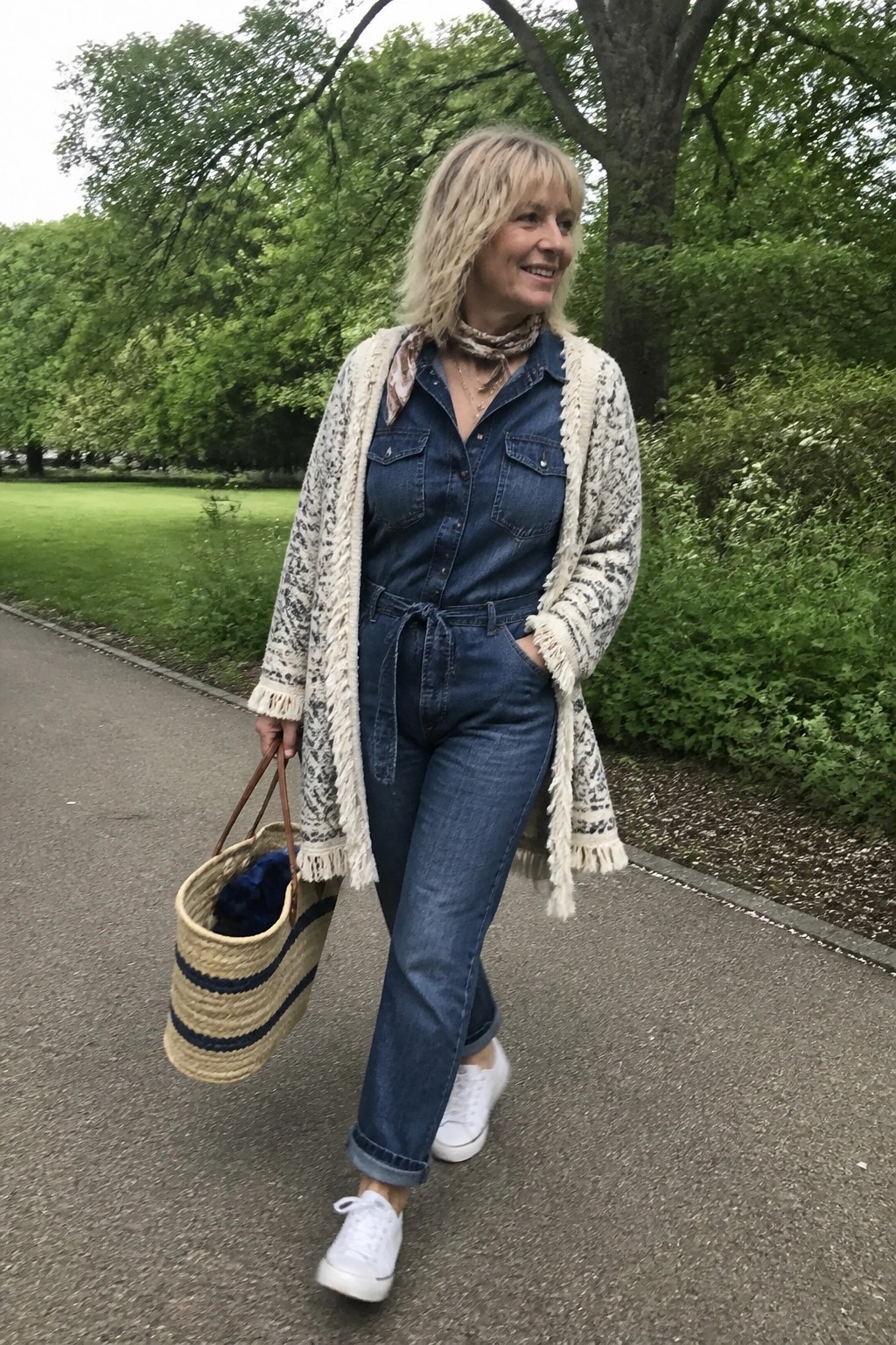 Blonde woman in denim jumpsuit and fringed cardigan carrying striped woven bag in park