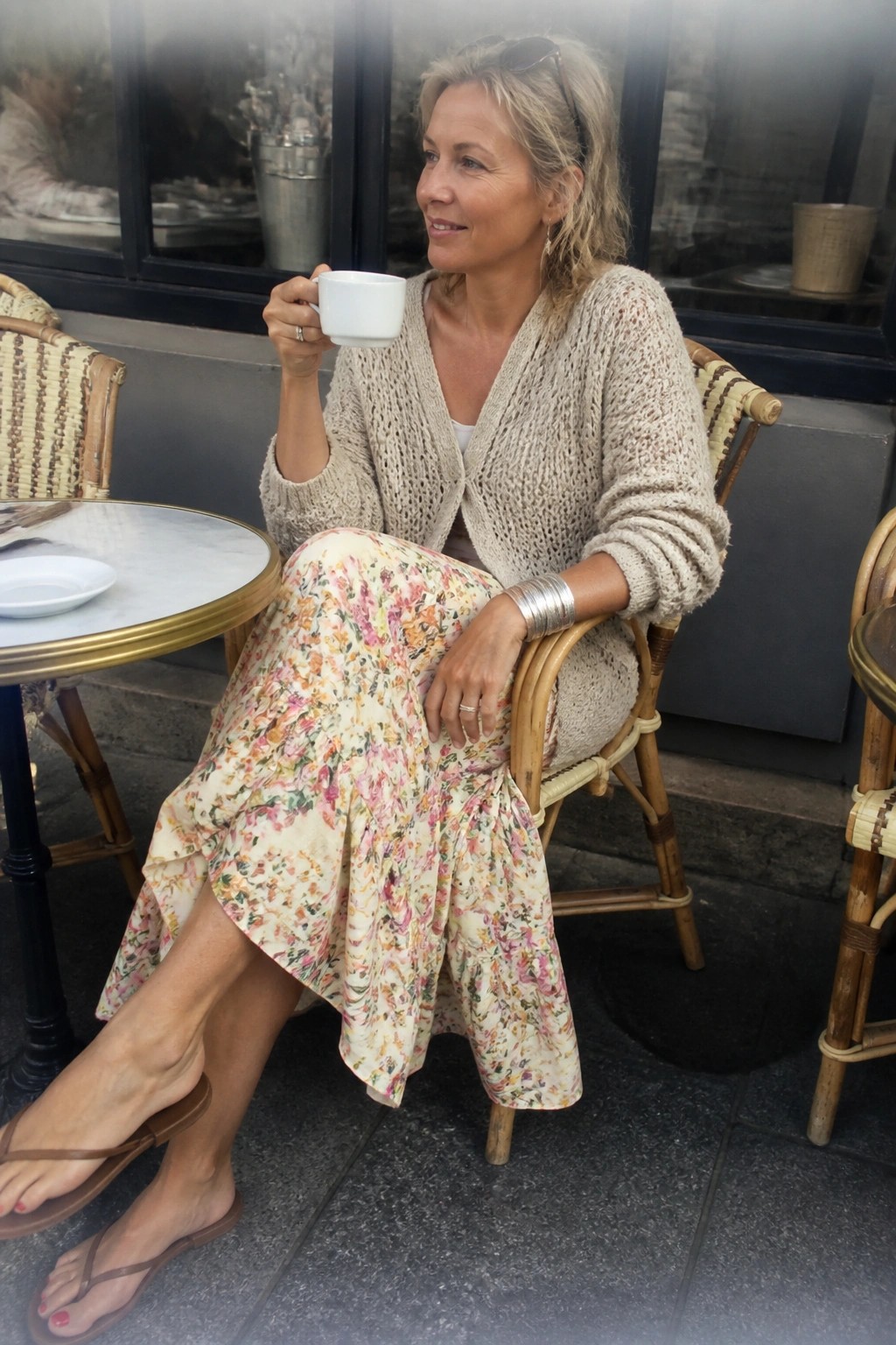 Woman in beige knit cardigan and floral skirt holding white cup at outdoor cafe