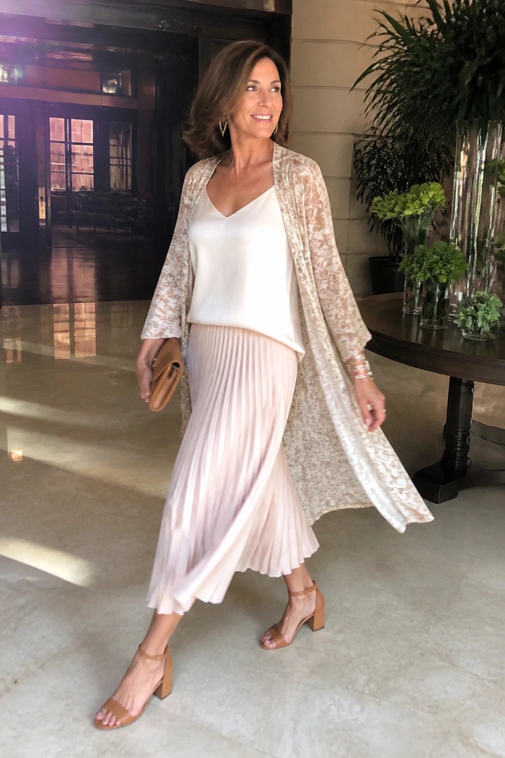 Woman in white top, pink pleated skirt, beige lace duster, holding clutch in lobby