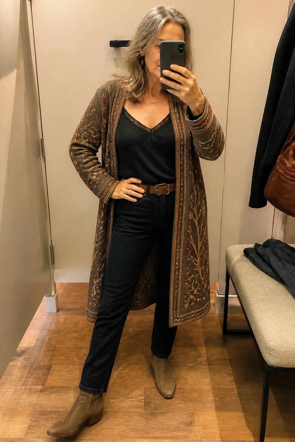 A woman over 50 wearing a long embroidered boho cardigan over a black lace-trim top and dark jeans with a brown belt and taupe suede ankle boots in a fitting room mirror selfie.