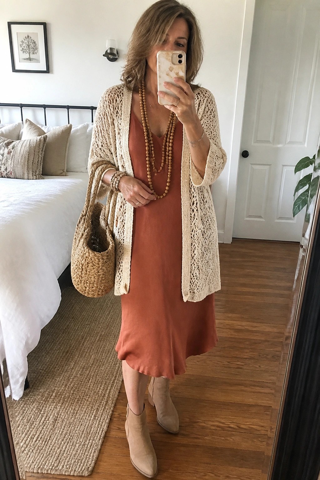 Woman in rust dress and cream open-knit cardigan taking bedroom mirror selfie with woven bag