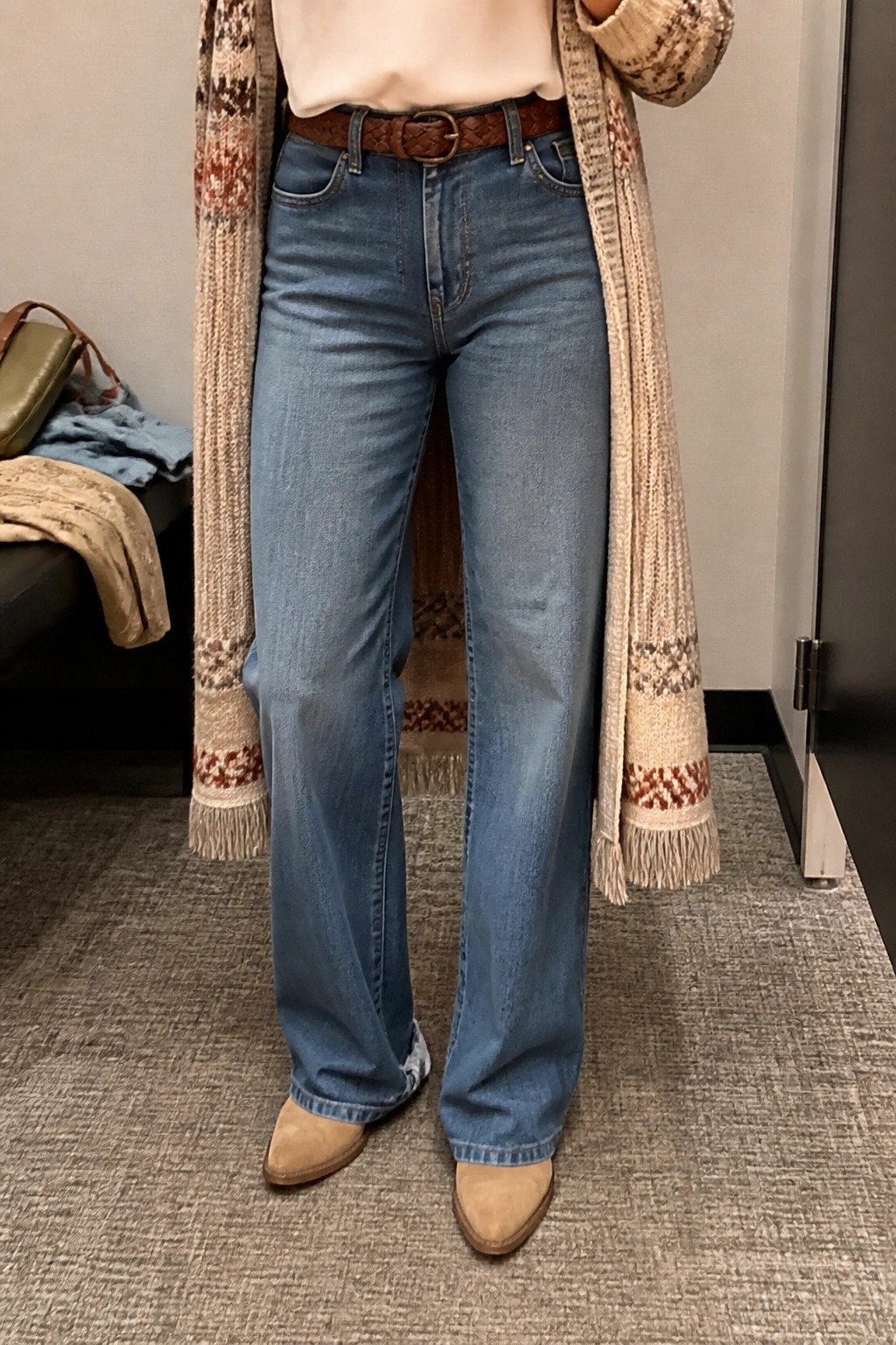 Person in wide-leg jeans, brown braided belt, fringed patterned cardigan, and beige boots