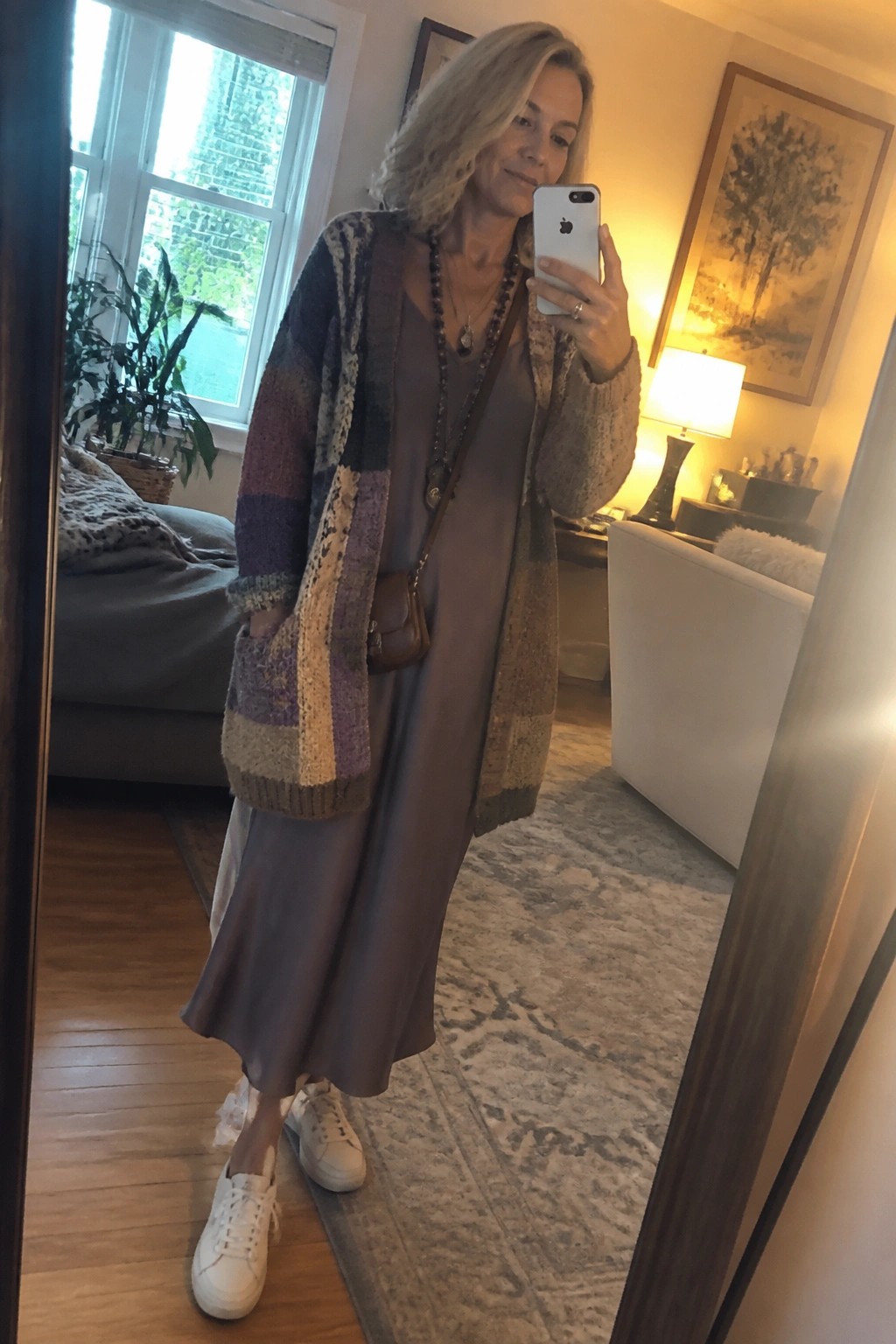 Woman over 50 in a multicolored patchwork cardigan layered over a taupe midi slip dress paired with white sneakers and beaded necklaces while holding a phone for a mirror selfie.