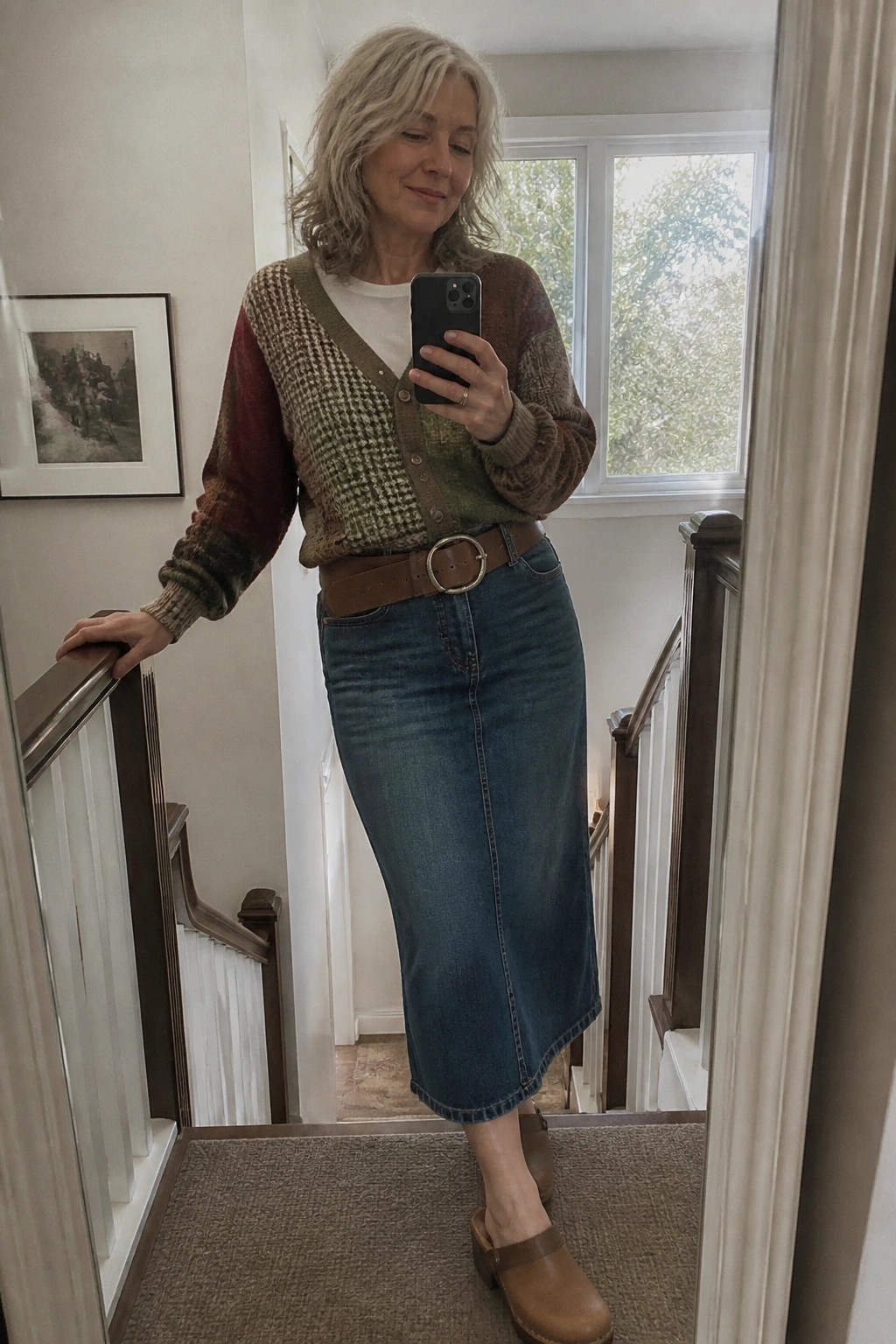 Gray-haired woman in patterned cardigan and denim skirt takes mirror selfie on stairs.