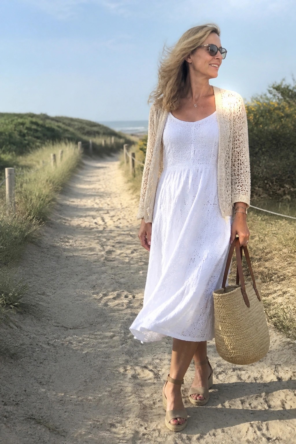Smiling woman in white eyelet dress and beige cardigan on beach path with straw bag.