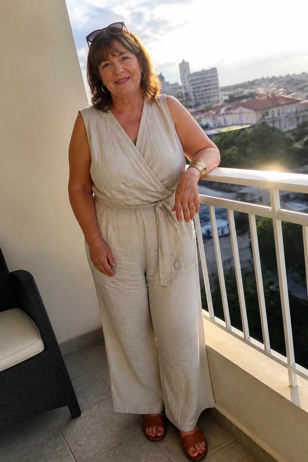 Woman in beige wrap jumpsuit leaning on sunny balcony railing overlooking city