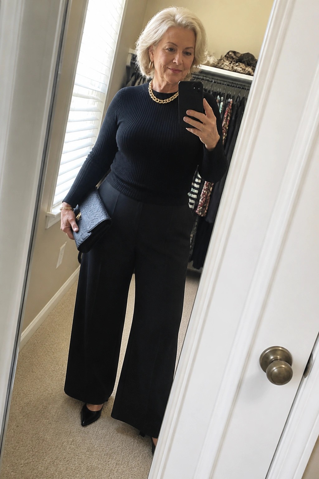 Woman with white hair in black ribbed sweater and wide pants holding smartphone and clutch