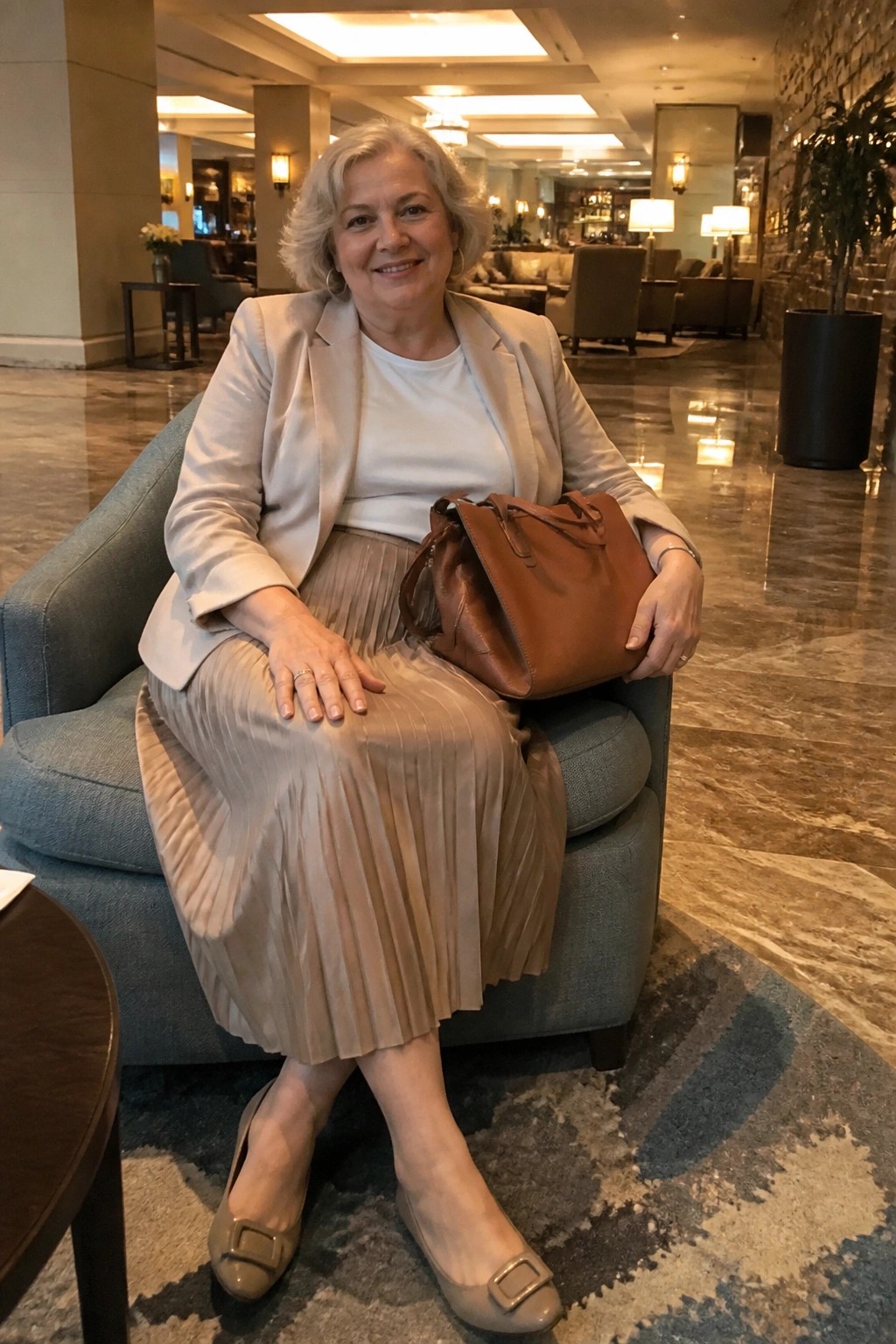 Gray-haired woman in beige blazer and pleated skirt holds brown bag in blue lobby armchair