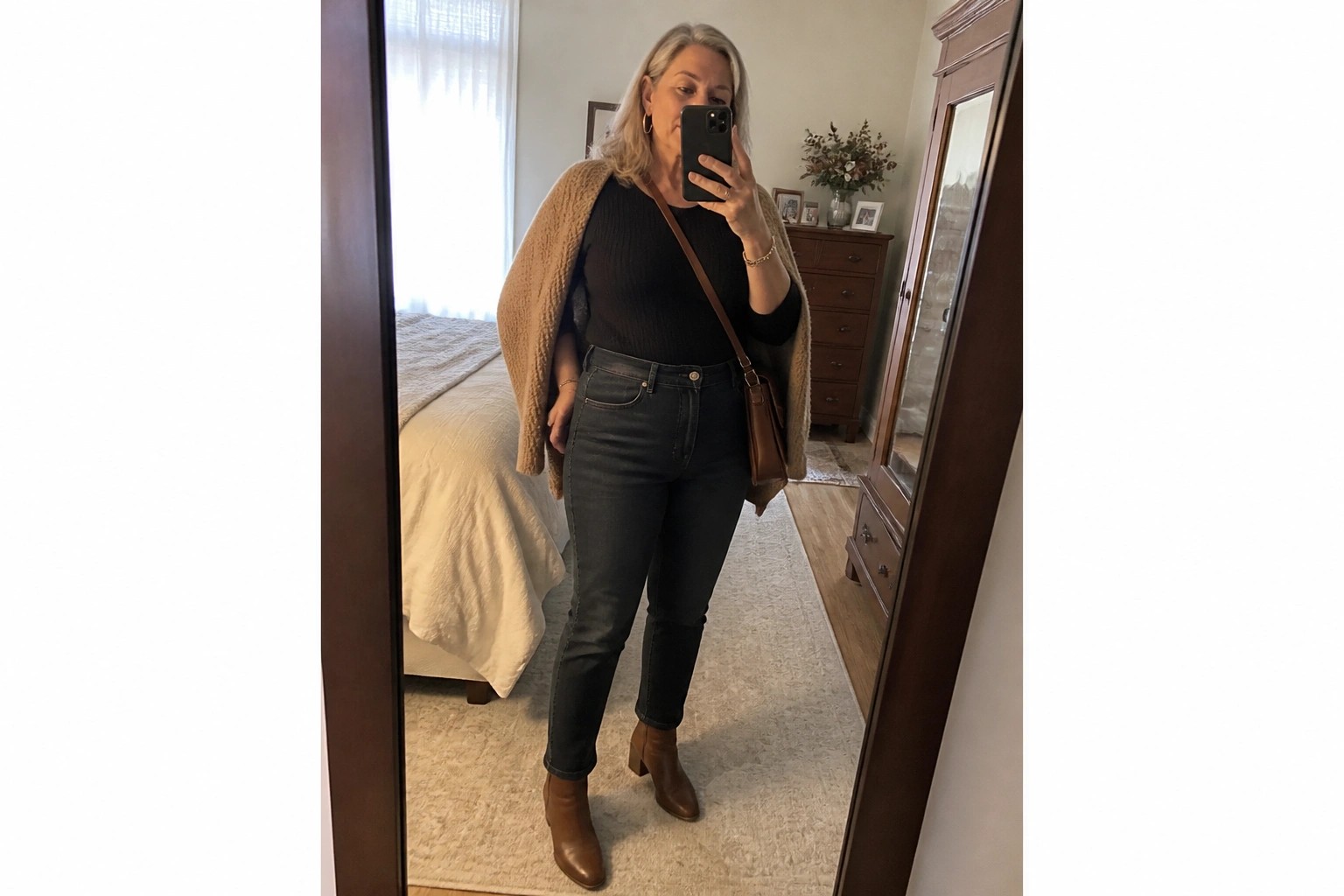 Woman in black top, dark jeans, tan cardigan taking mirror selfie in bedroom