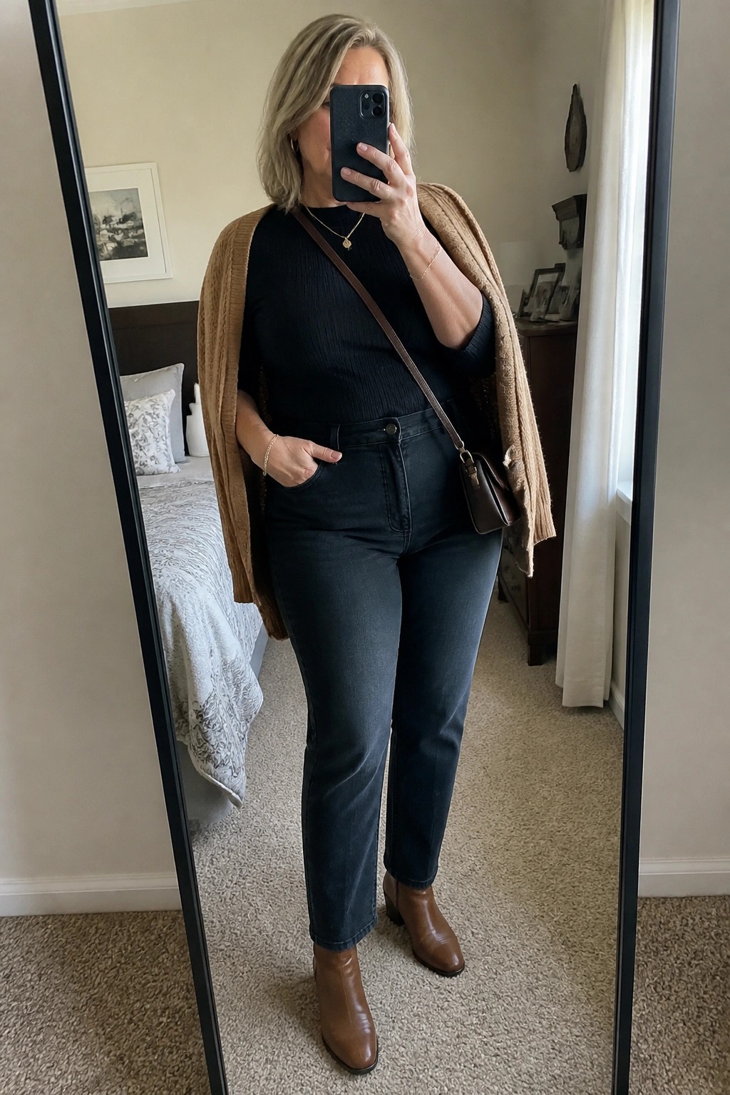 Woman in black top, dark jeans, tan cardigan taking mirror selfie in bedroom