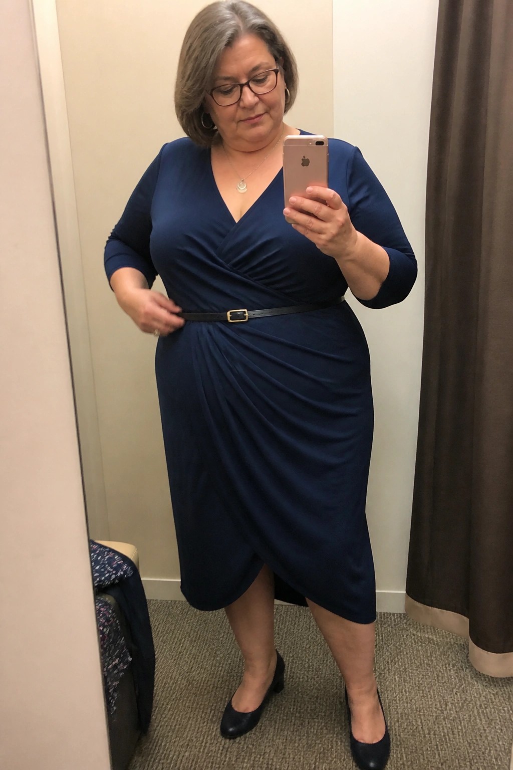 Woman with gray hair in navy wrap dress takes mirror selfie holding iPhone