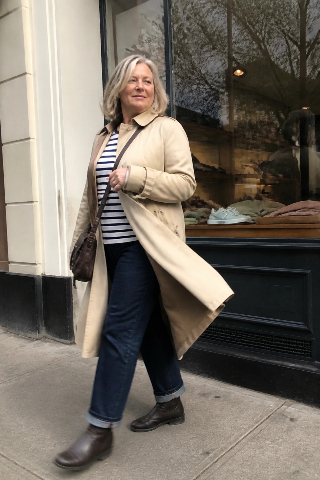 Woman over 50 in plus size outfit wearing open beige trench coat over blue and white striped shirt with dark blue jeans cuffed at the ankle brown leather boots and brown crossbody bag standing on sidewalk.