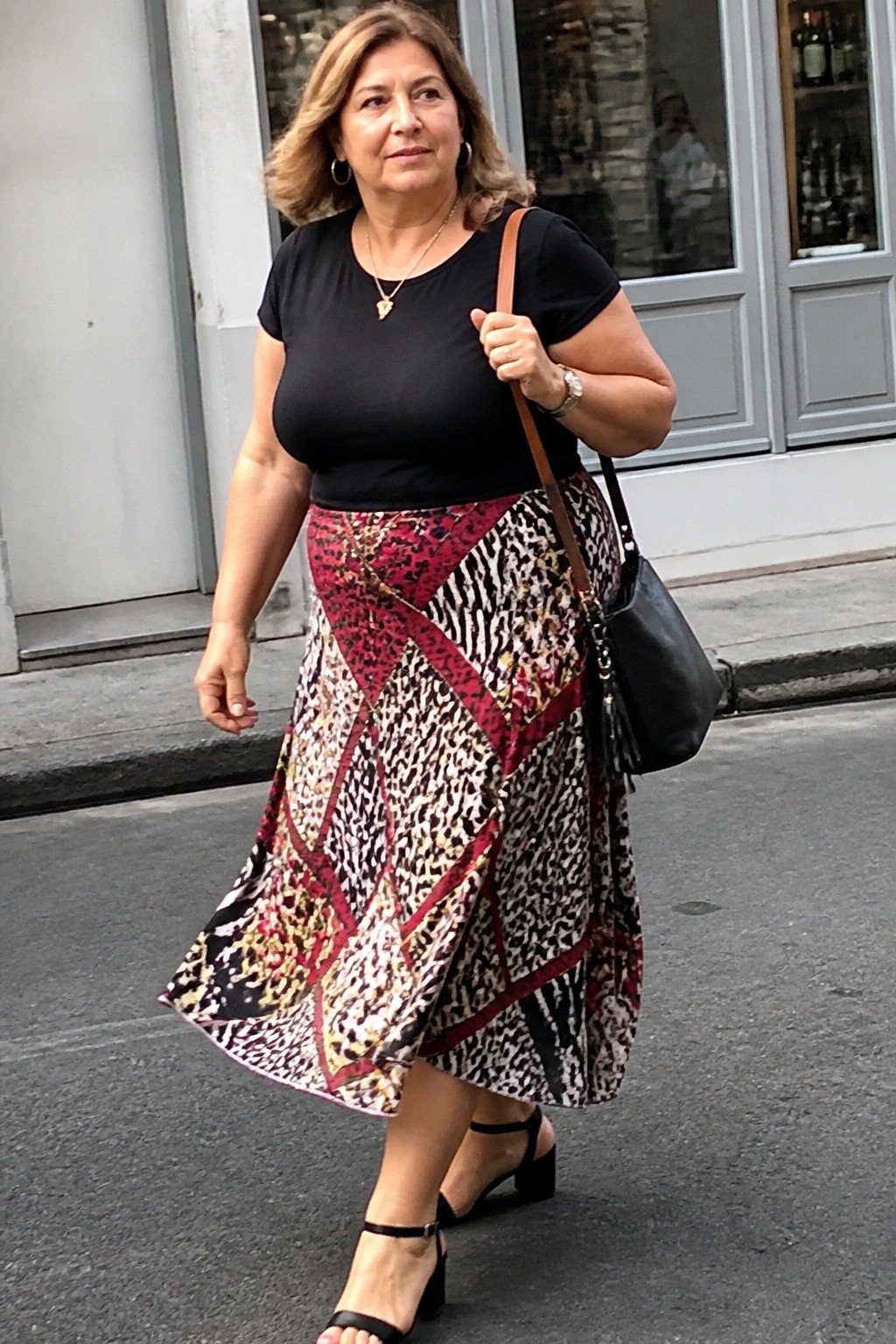 Woman in black top and red-black animal-print skirt walking with black handbag.