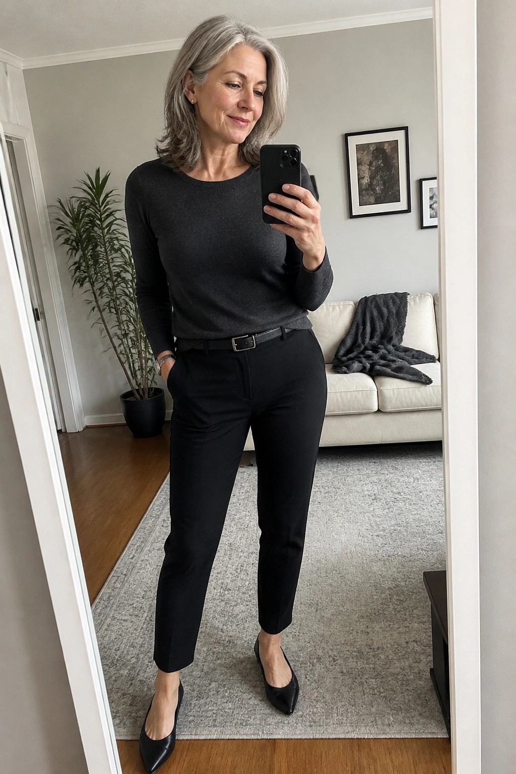 Gray-haired woman in black sweater and pants taking mirror selfie in living room