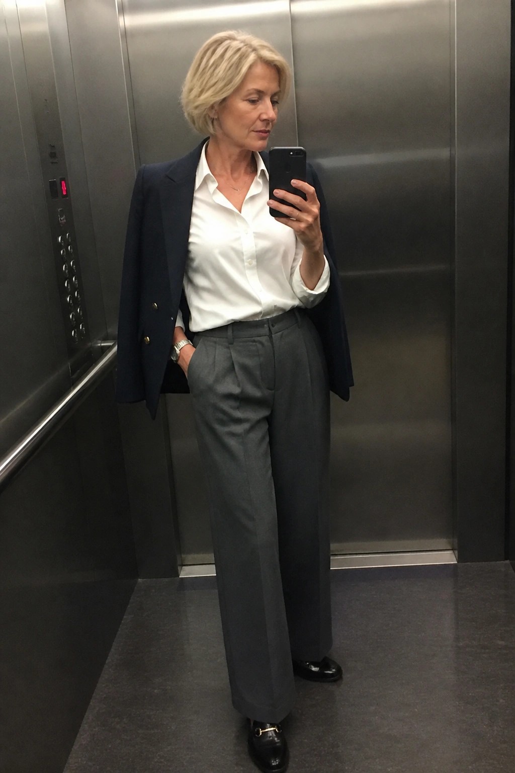 Blonde woman in navy blazer, white shirt, and gray trousers taking phone selfie in elevator.